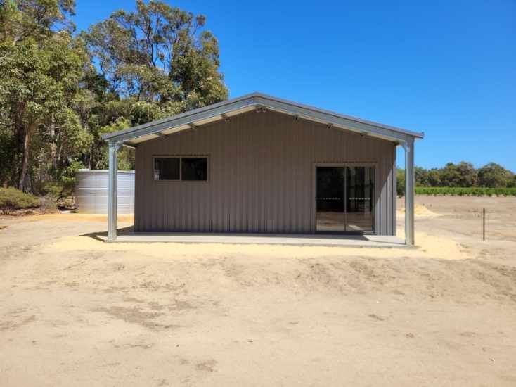 A brown metal shed with a sliding glass door and window sits on a sandy lot near trees and a water tank.