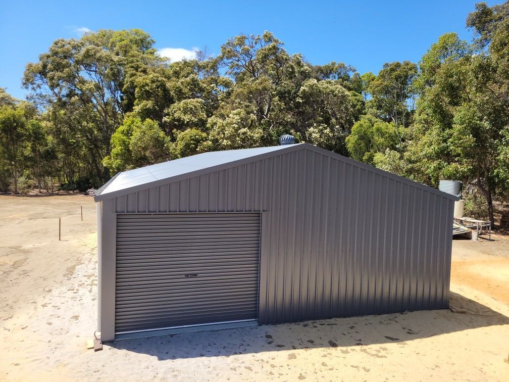 A grey metal shed with a roller door, situated on a cleared dirt lot in front of a line of green trees under a blue sky.