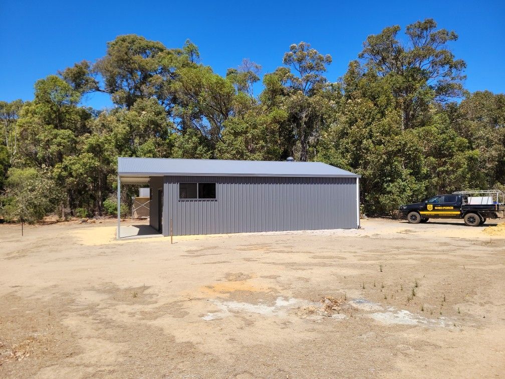A gray corrugated metal shed with a side porch sits on a patch of dirt in a forest clearing next to a dark pickup truck.