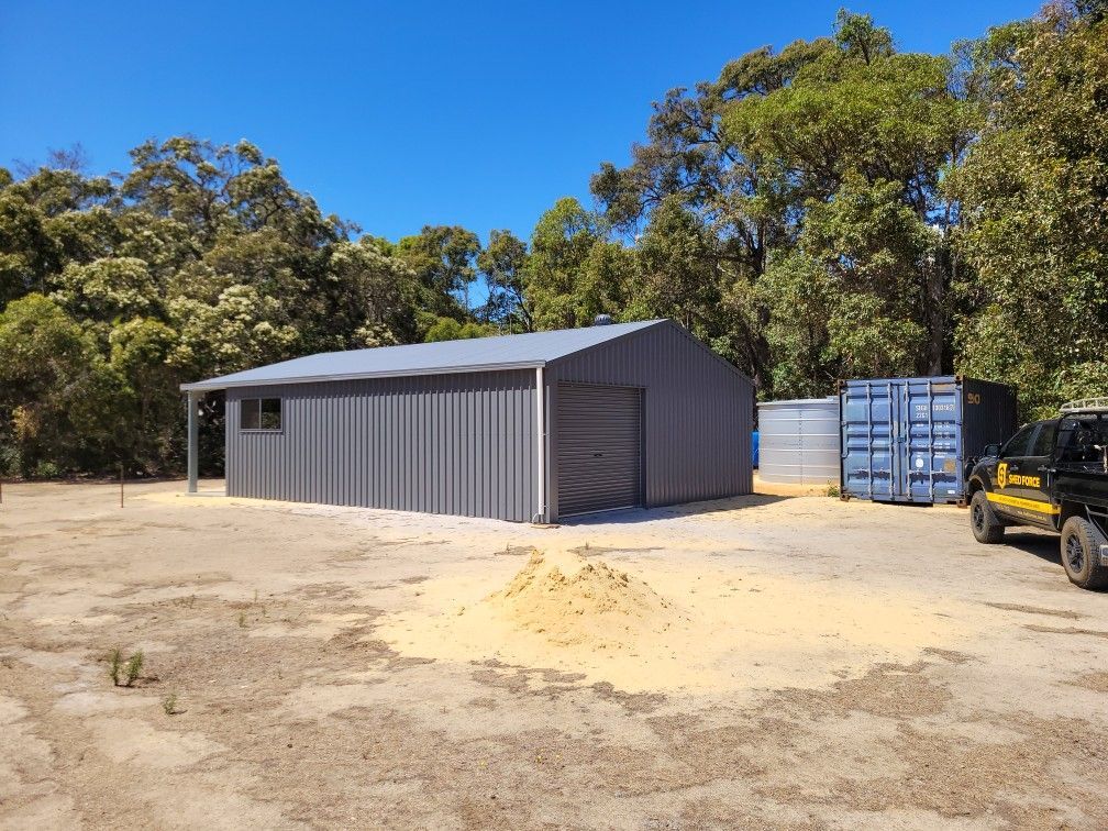 A gray metal shed sits in a clearing surrounded by trees, with a blue shipping container and a parked truck nearby.