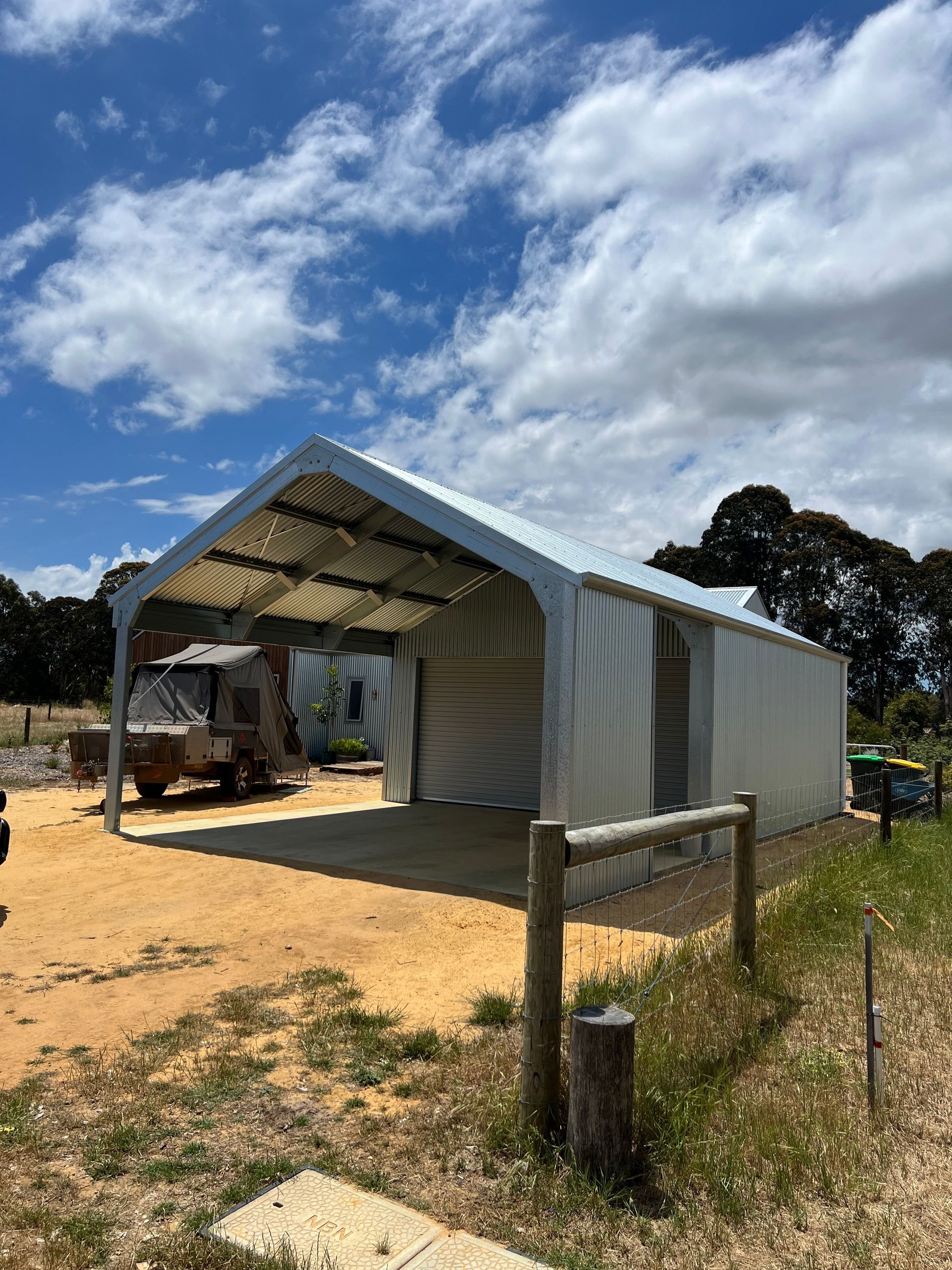 A white fabric-covered metal carport structure standing on a dirt lot under a blue sky with clouds.