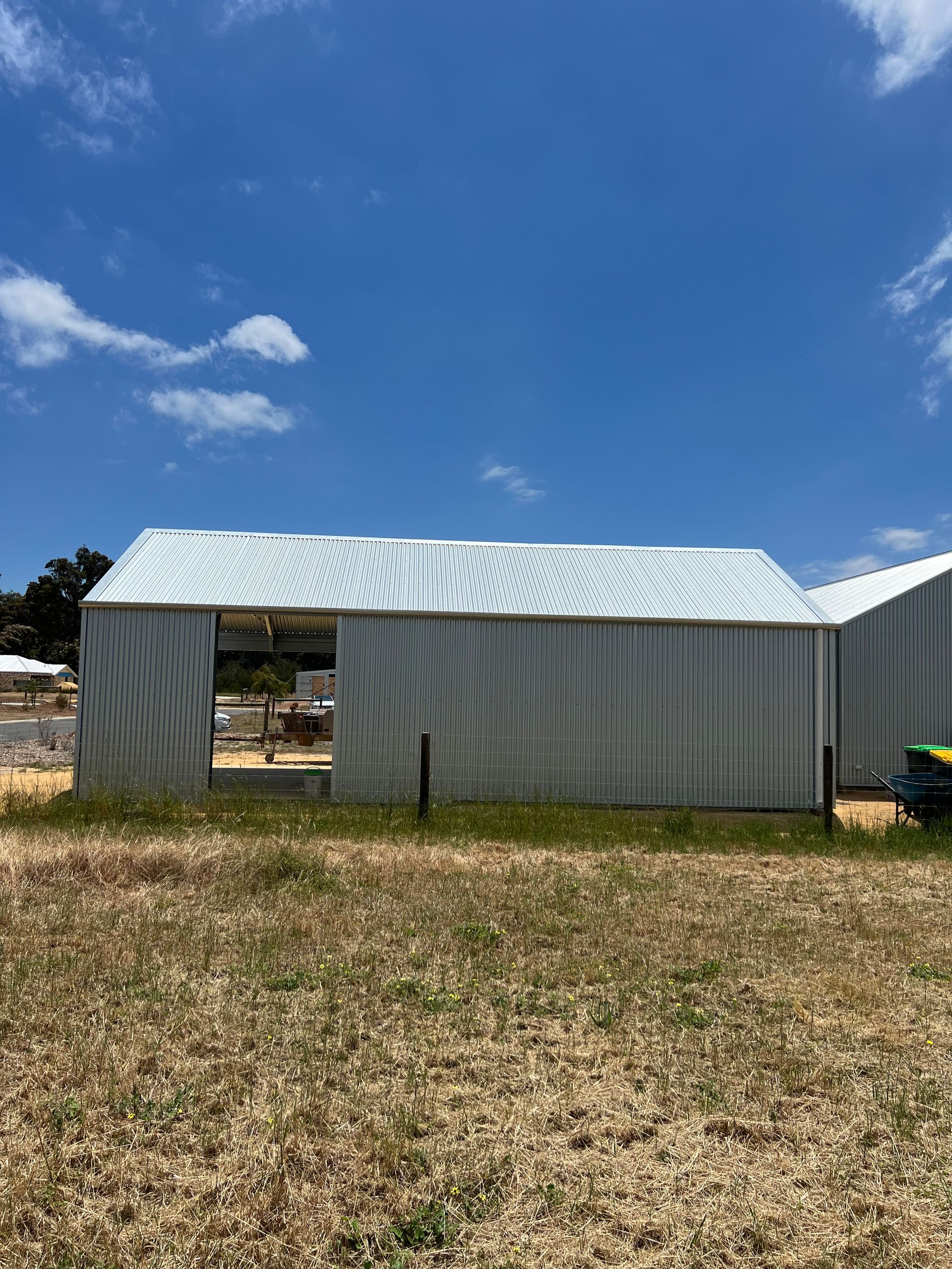 A silver corrugated metal shed under construction, standing in a dry, grassy field against a bright blue sky.