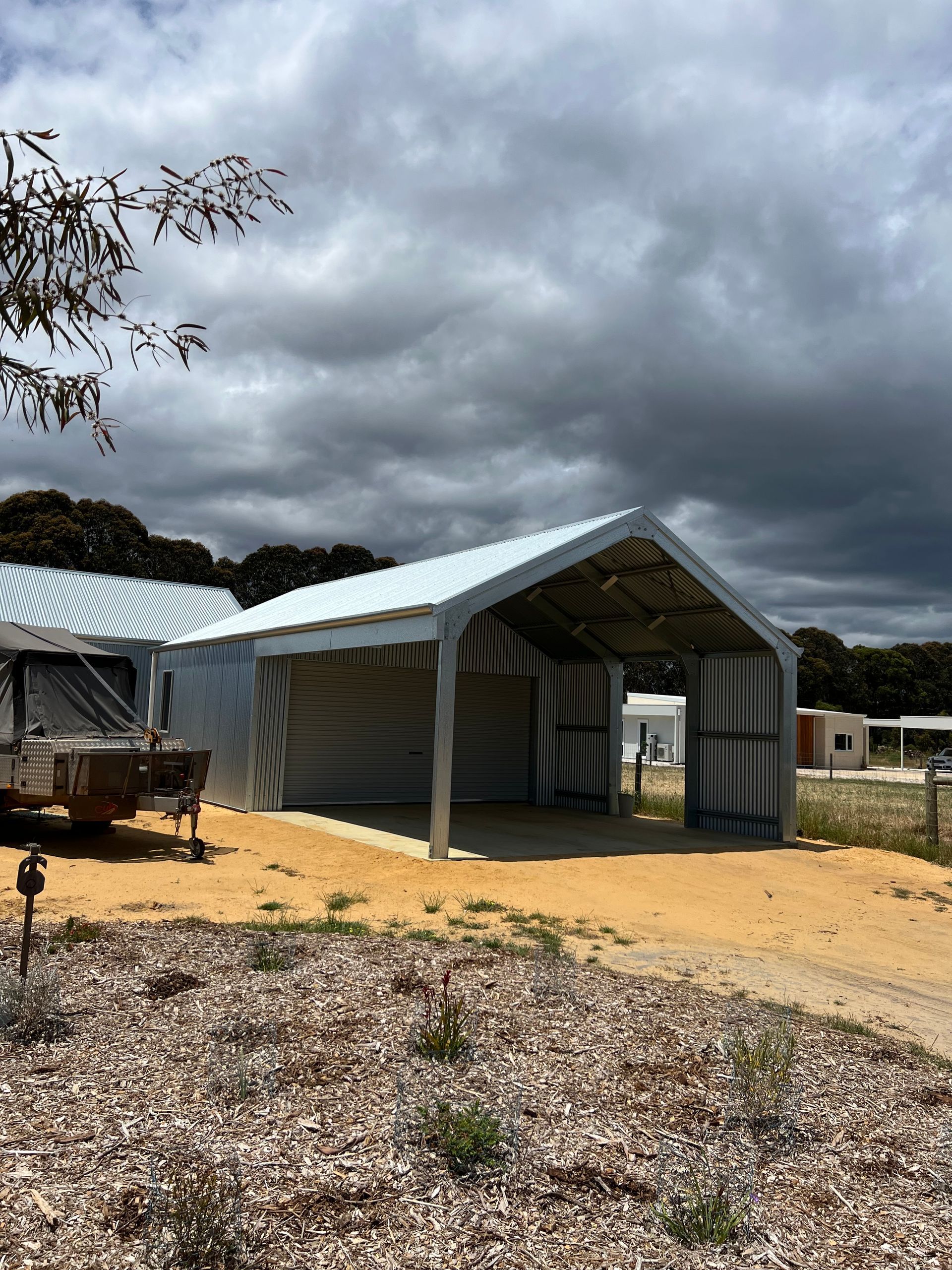 A metal-framed, open-front carport with a gabled roof standing on a dirt lot under a cloudy sky.
