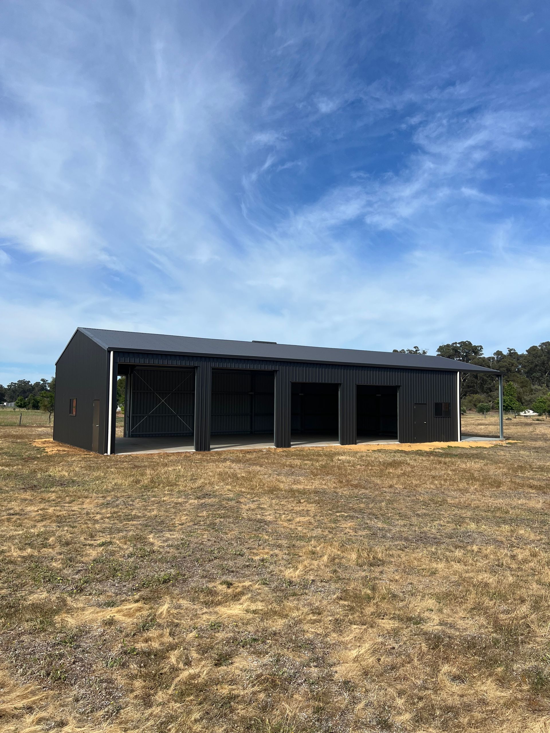 A dark grey, rectangular open-fronted metal shed sits in a dry, grassy field under a bright blue sky with light clouds.