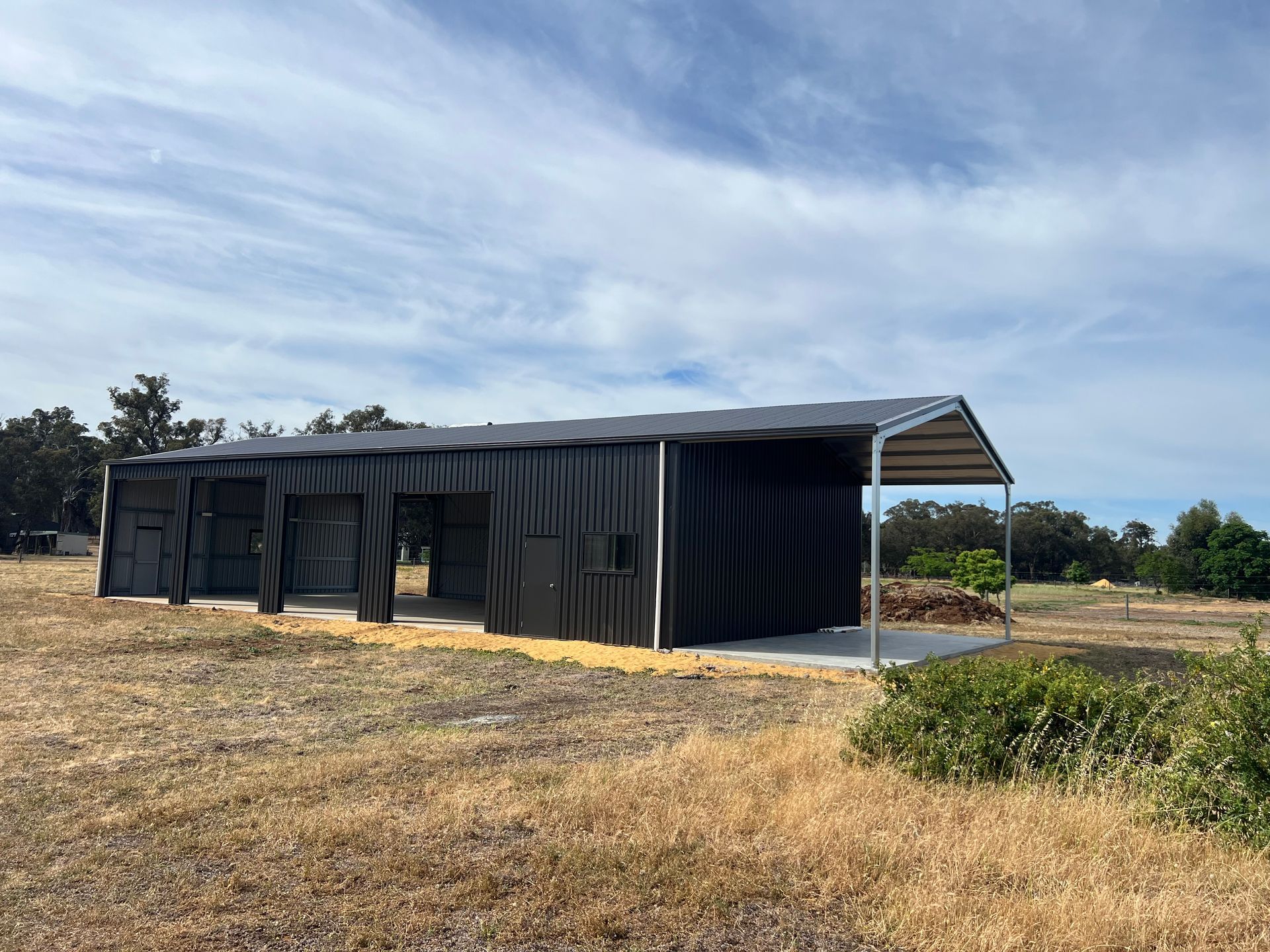 A black, industrial-style shed with an open-front carport area, situated in a dry, grassy field under a cloudy sky.