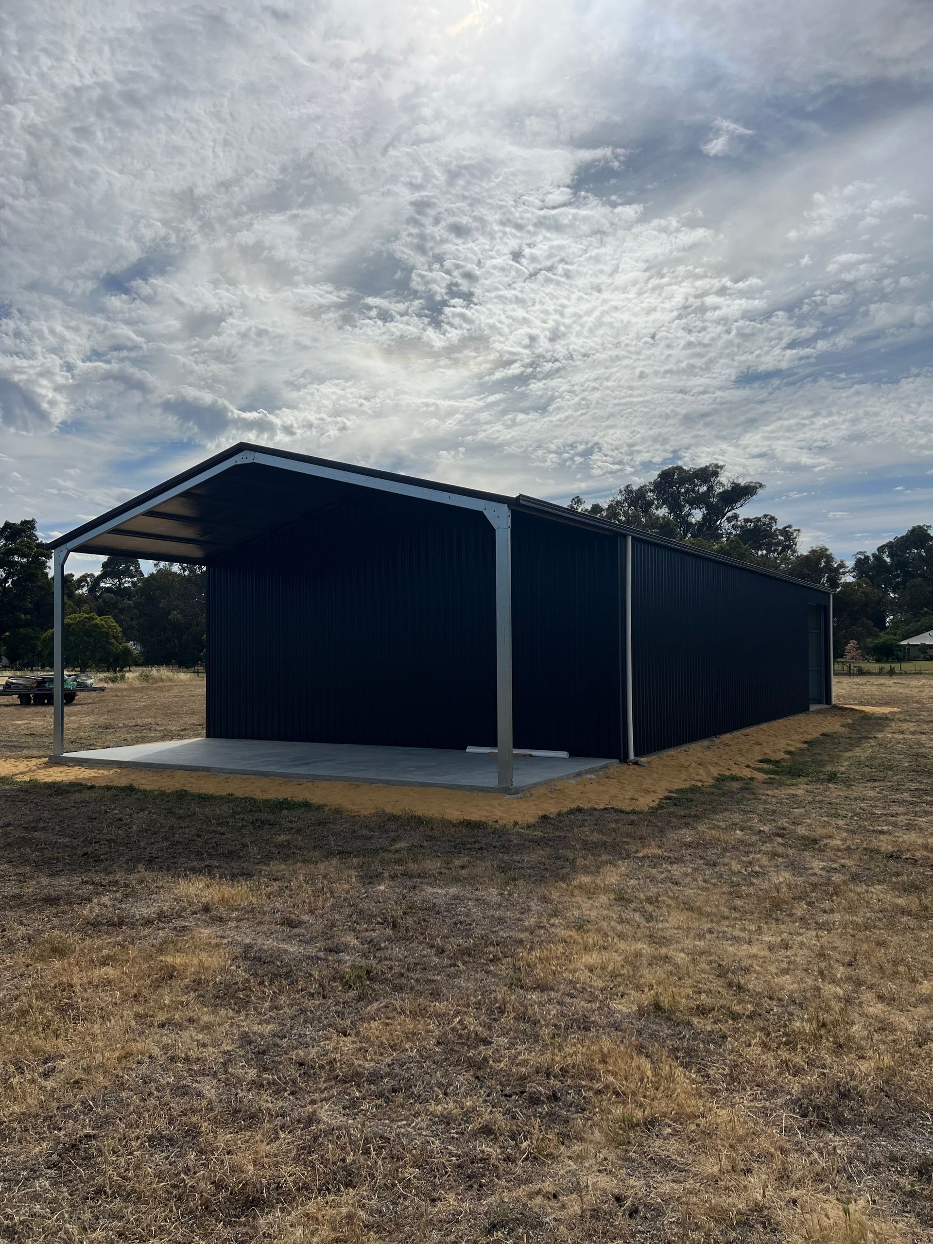 A black metal shed with an open front and a concrete base sits on a dry, grassy plot under a cloudy sky.