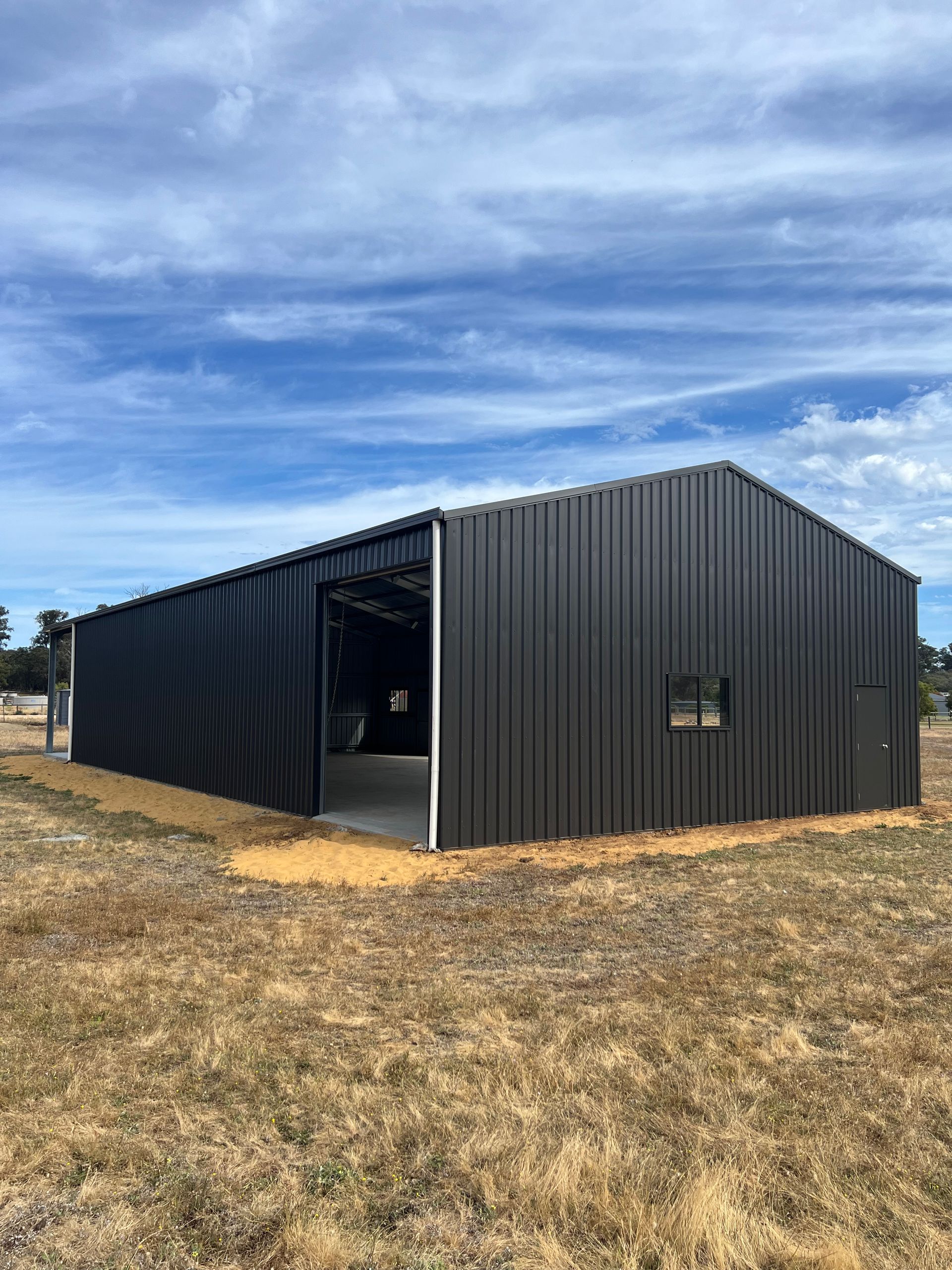 A rectangular, dark-gray corrugated metal building stands in a dry, grassy field under a blue, cloudy sky.