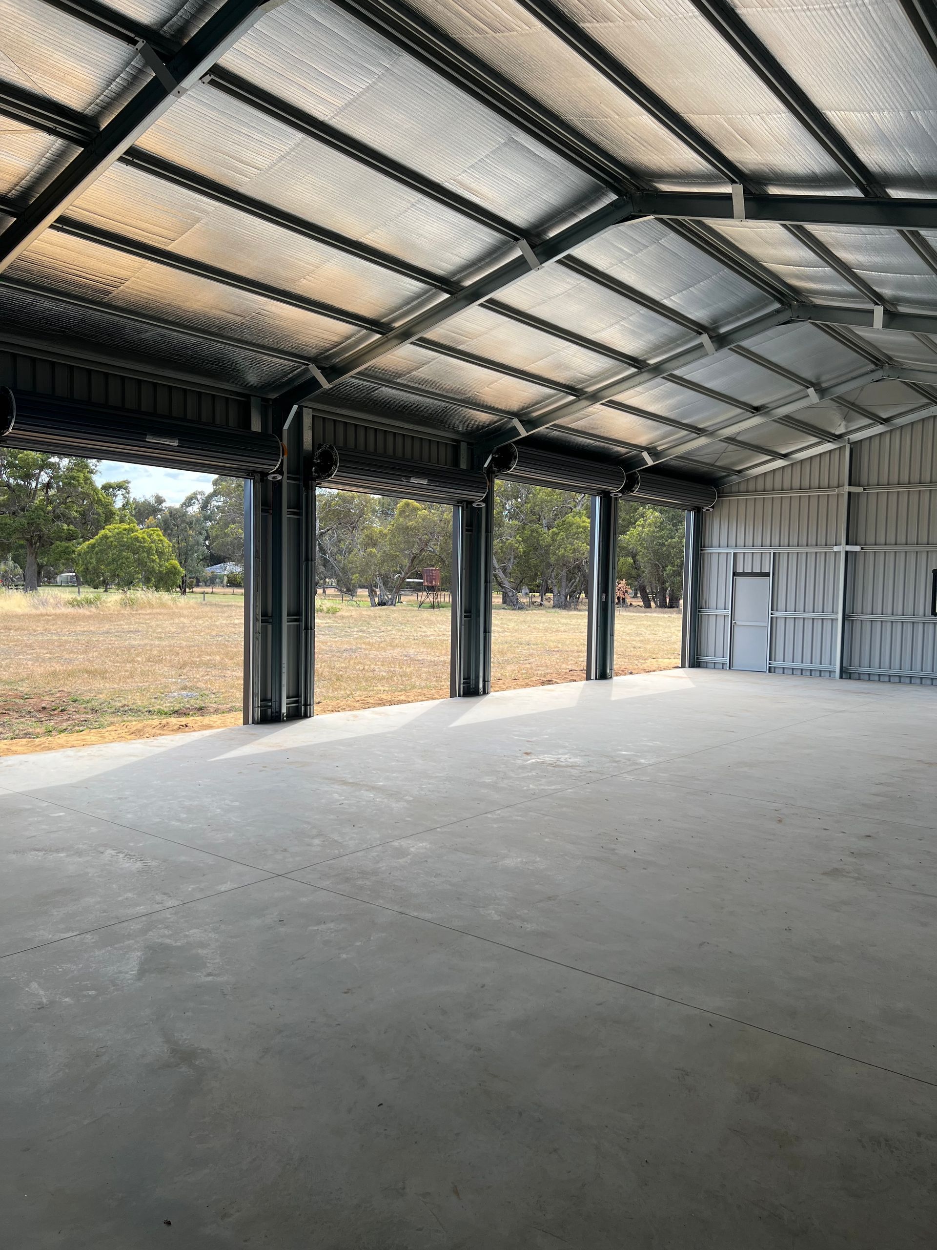 An empty, open-sided steel shed with a concrete floor, metal frame, and silver insulated roof, overlooking an open field.