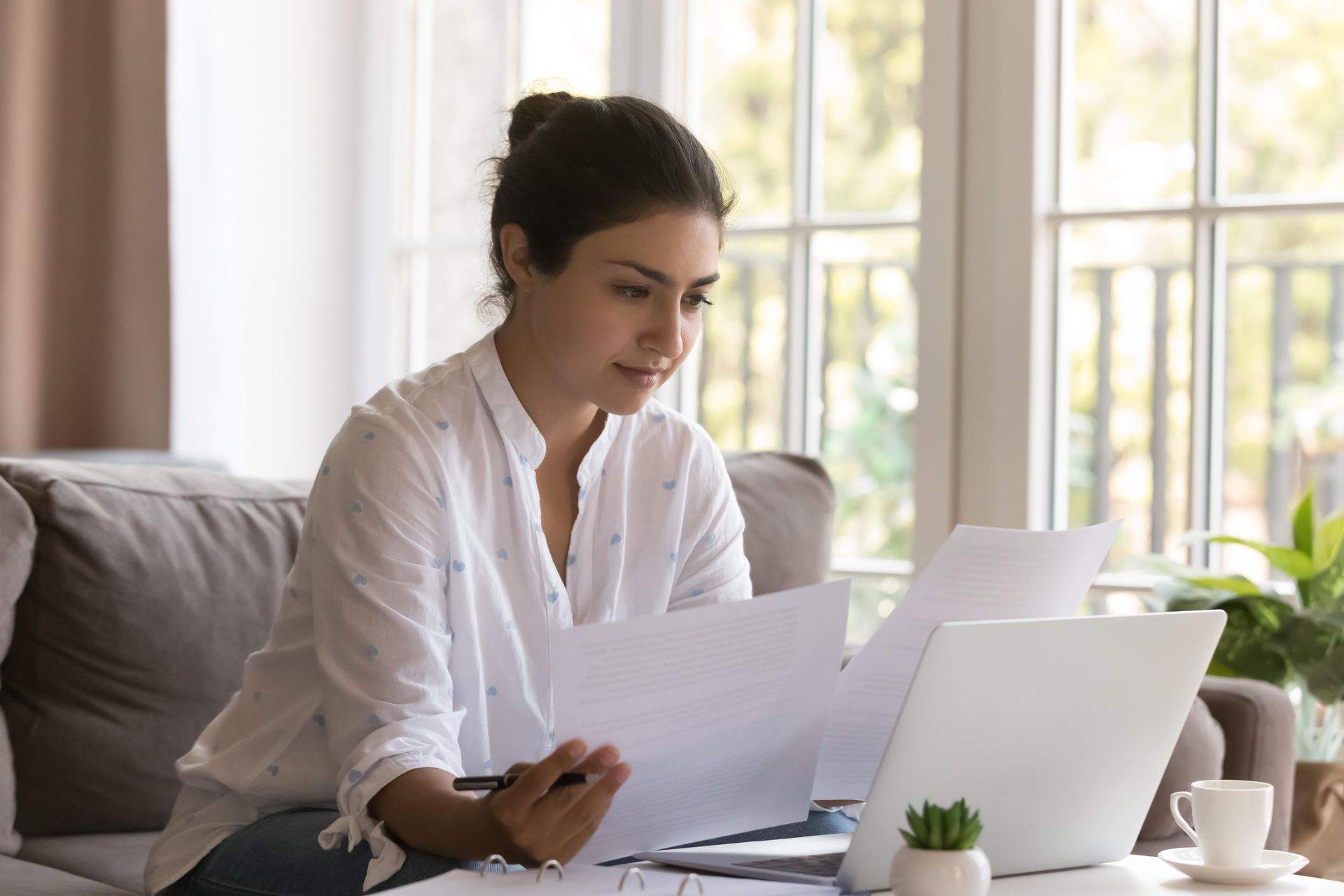 A woman is sitting on a couch looking at papers and a laptop.