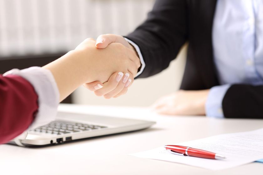 Two people are shaking hands over a table with a laptop.