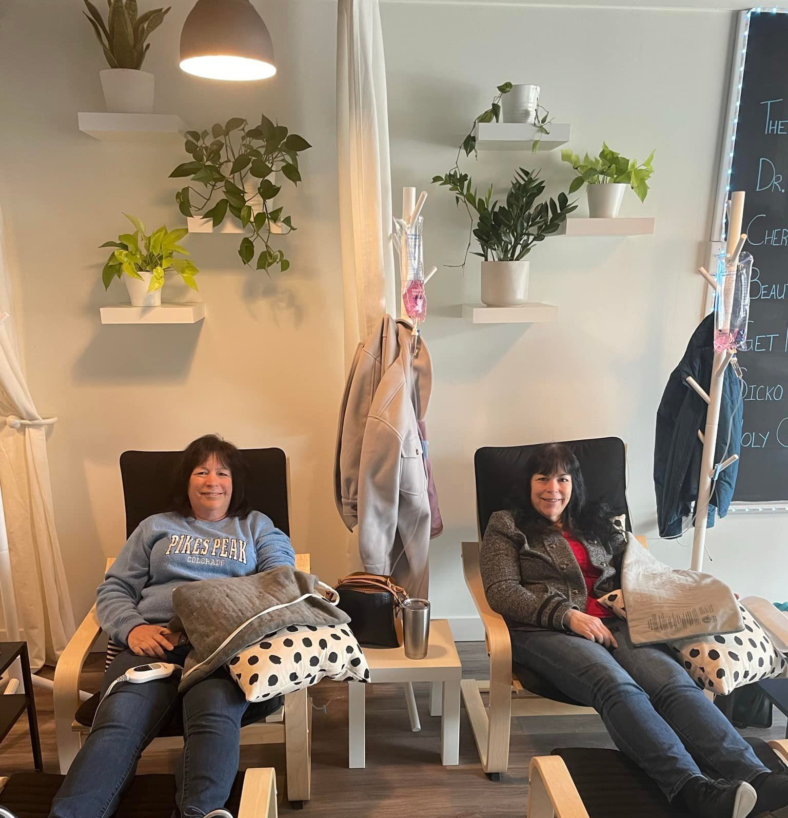 Two women are sitting in chairs in a room with plants on shelves