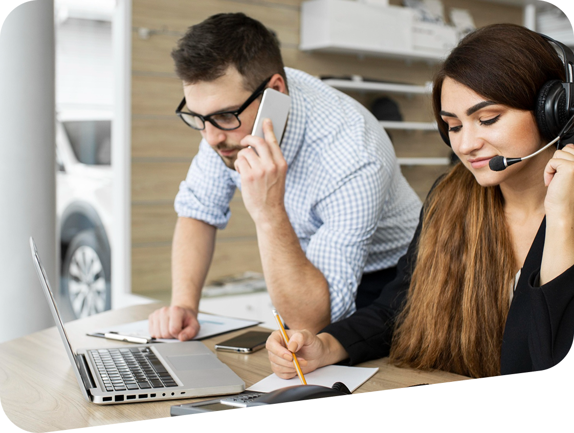 A man and a woman are sitting at a desk with a laptop and a phone.