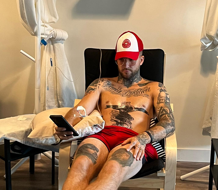 A shirtless man with tattoos is sitting in a chair holding a cell phone.