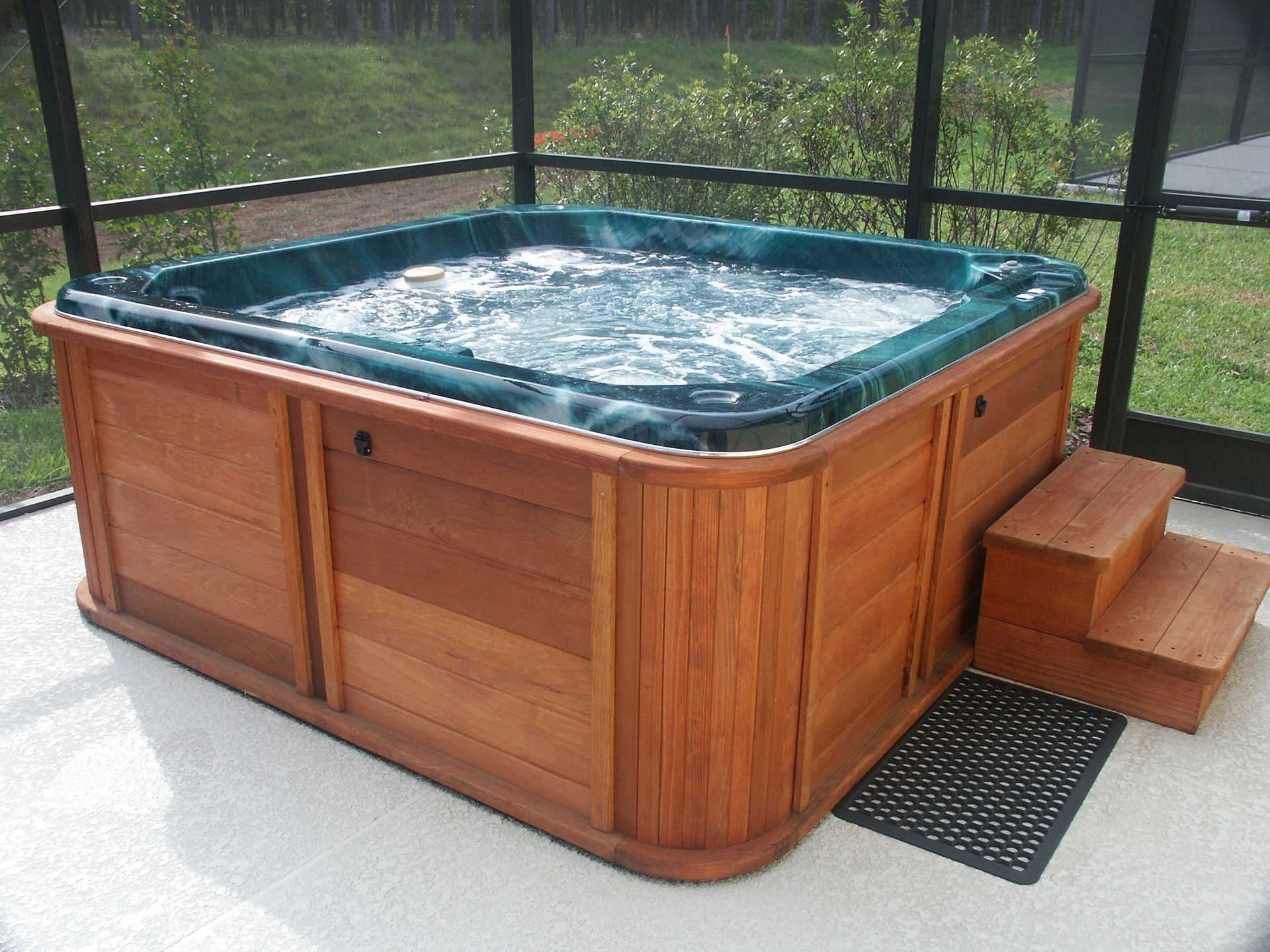 A wooden hot tub on a concrete deck is filled with water and walled by screens.