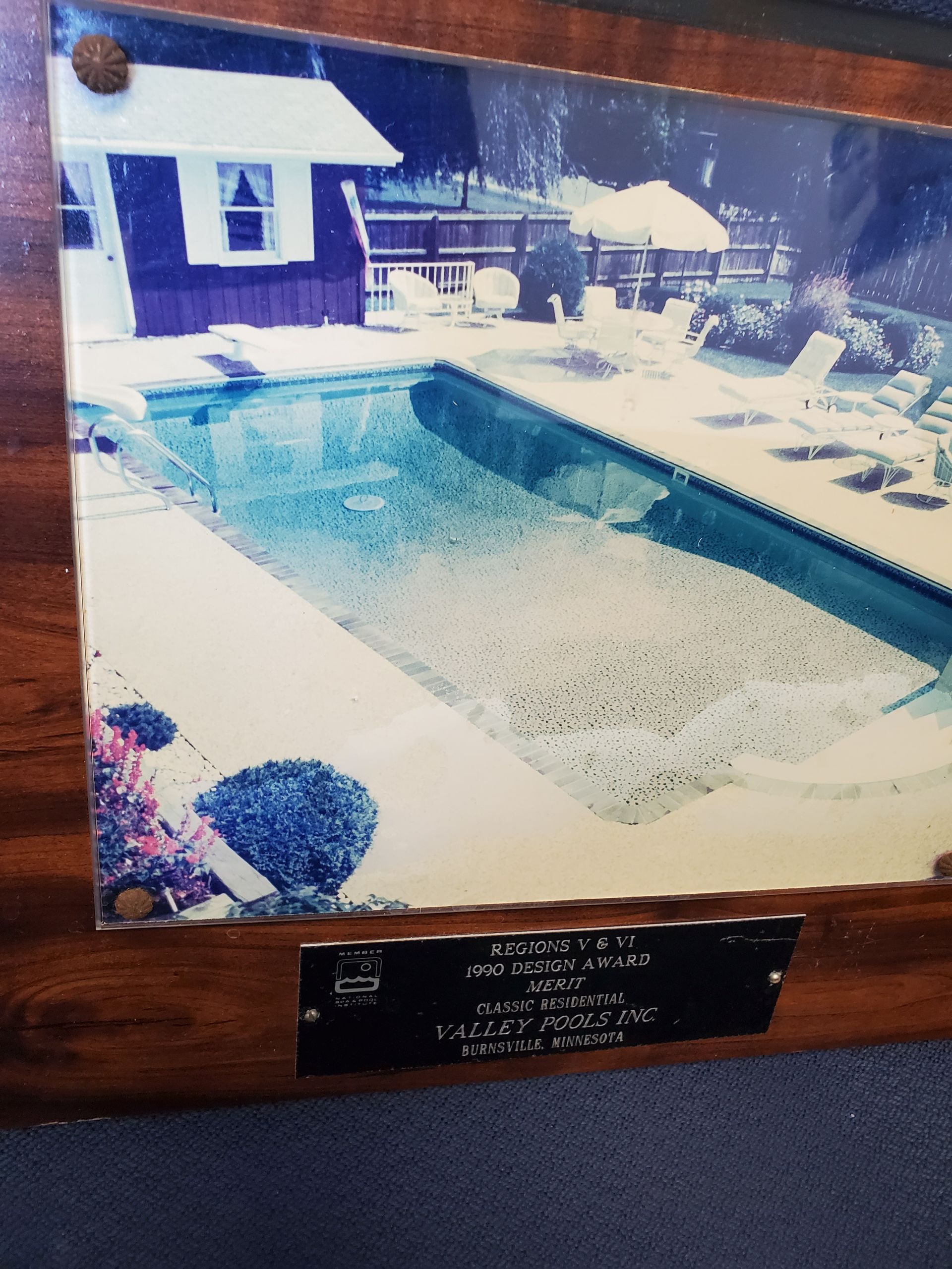 Framed photo of a rectangular pool with a blue house to the left. Brown wooden frame and a plaque below the image.