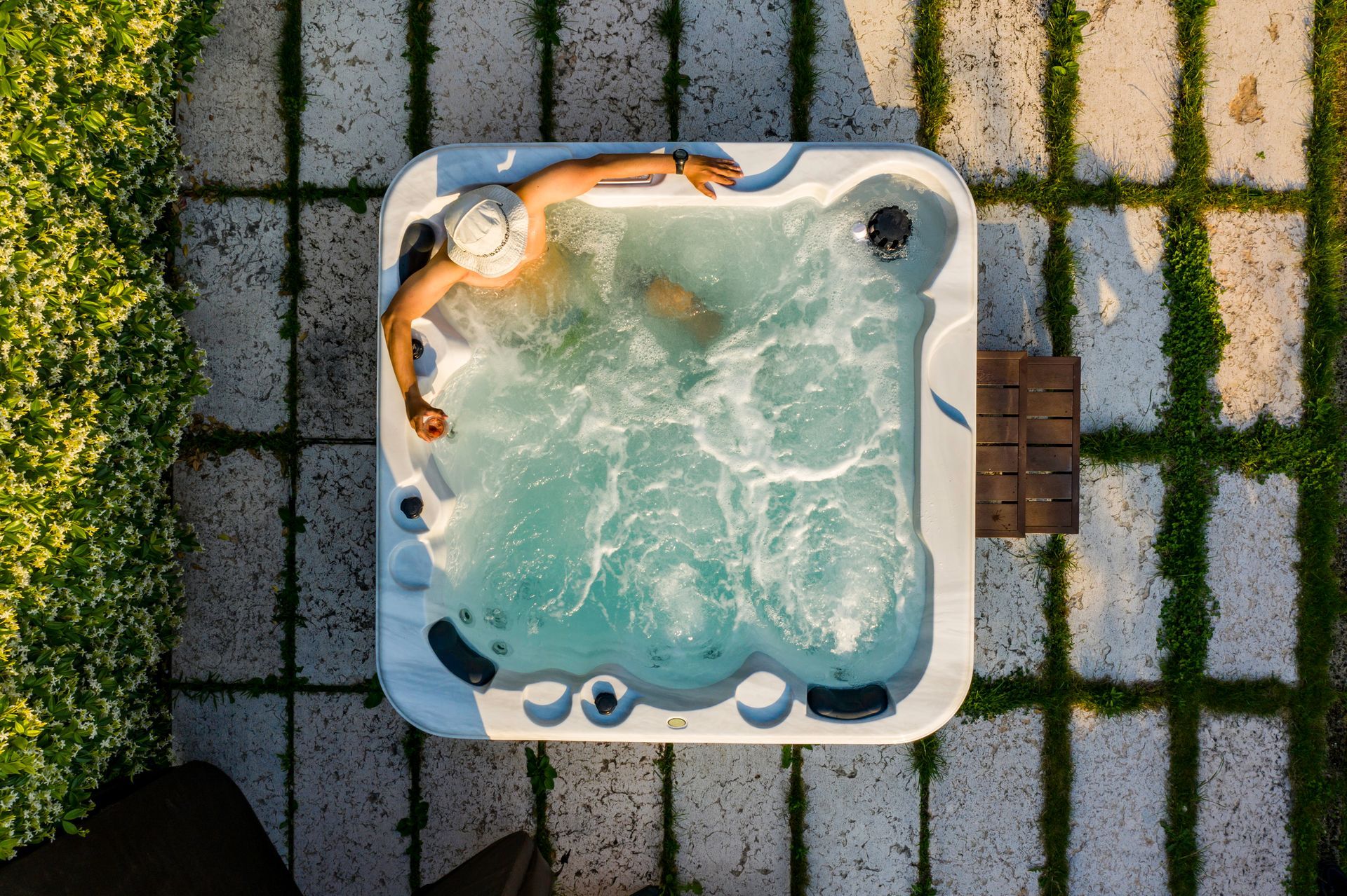 Person relaxes in hot tub surrounded by greenery and stone tiles.