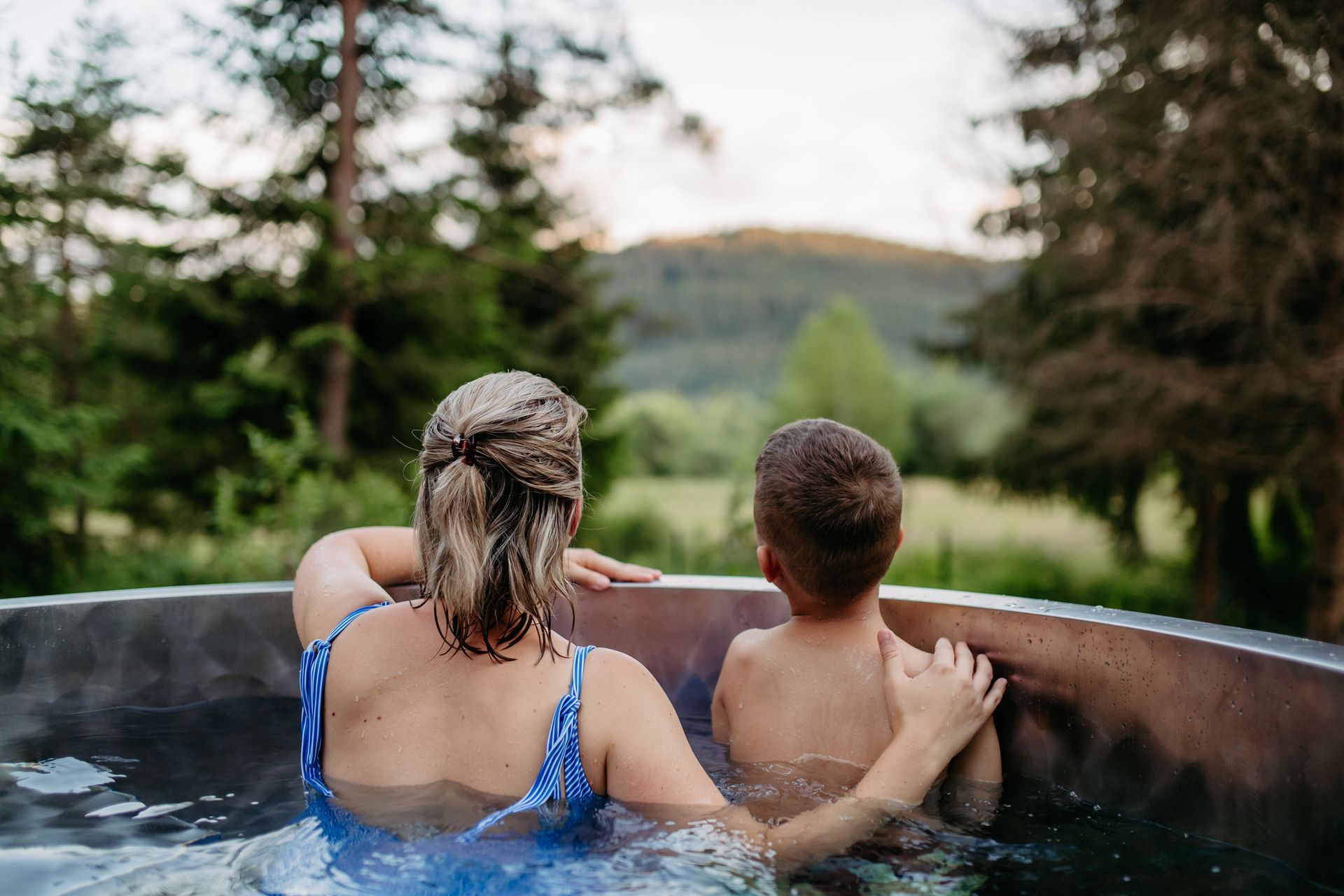Women and kid relax in hot tub with lake and forest view at sunset.