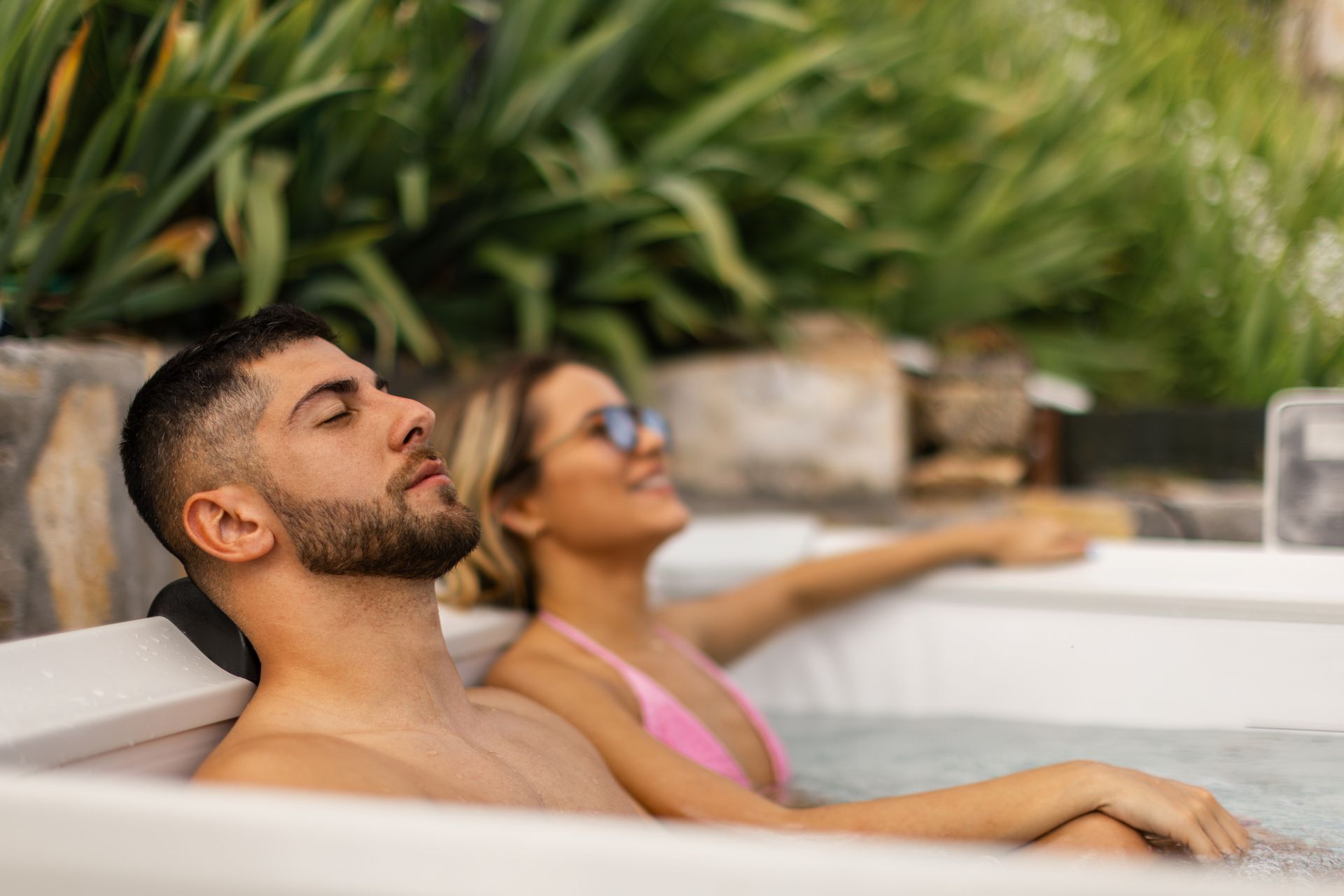 Two people relaxing in an outdoor hot tub surrounded by greenery and stone features.
