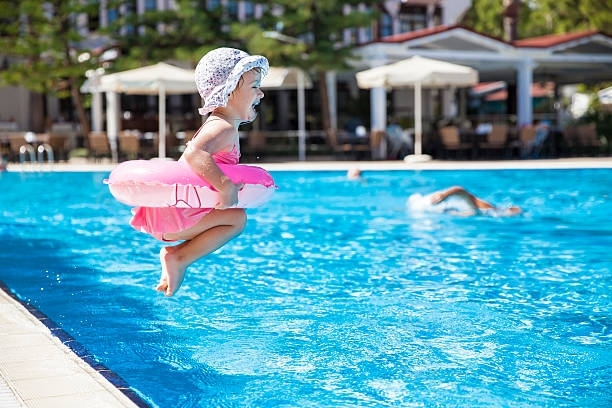 Little girl dressed in a pink swimsuit and wearing a safety ring jumps into a pool while smiling.