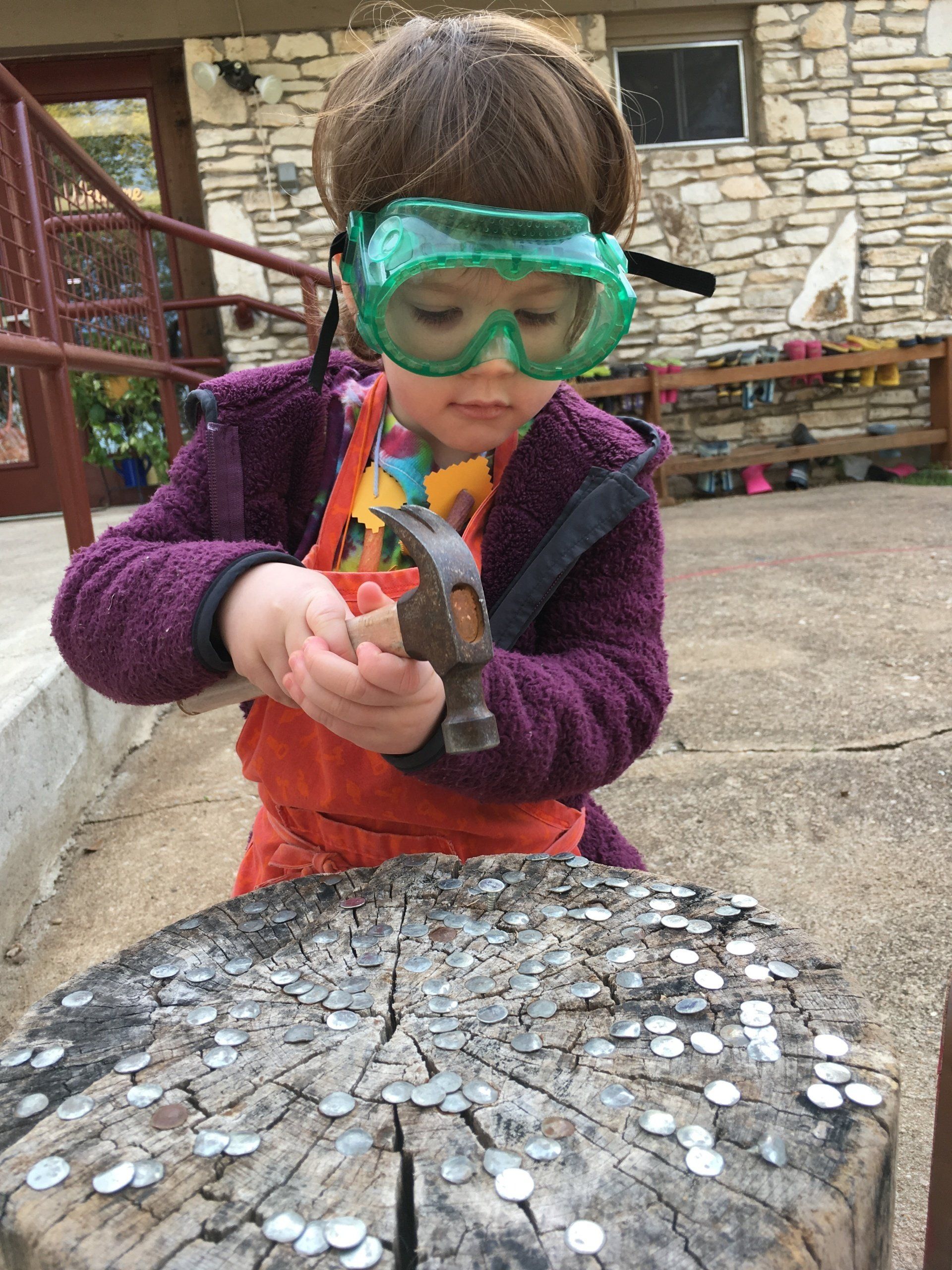A little girl wearing goggles is hammering nails into a tree stump.