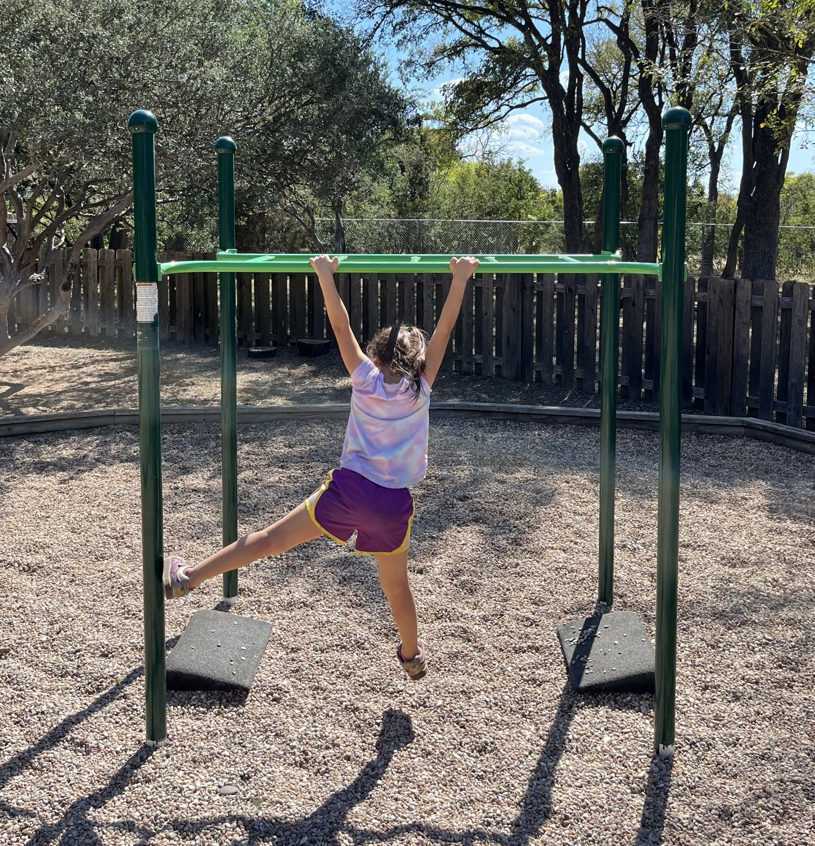 A young girl is hanging on a green bar in a playground.