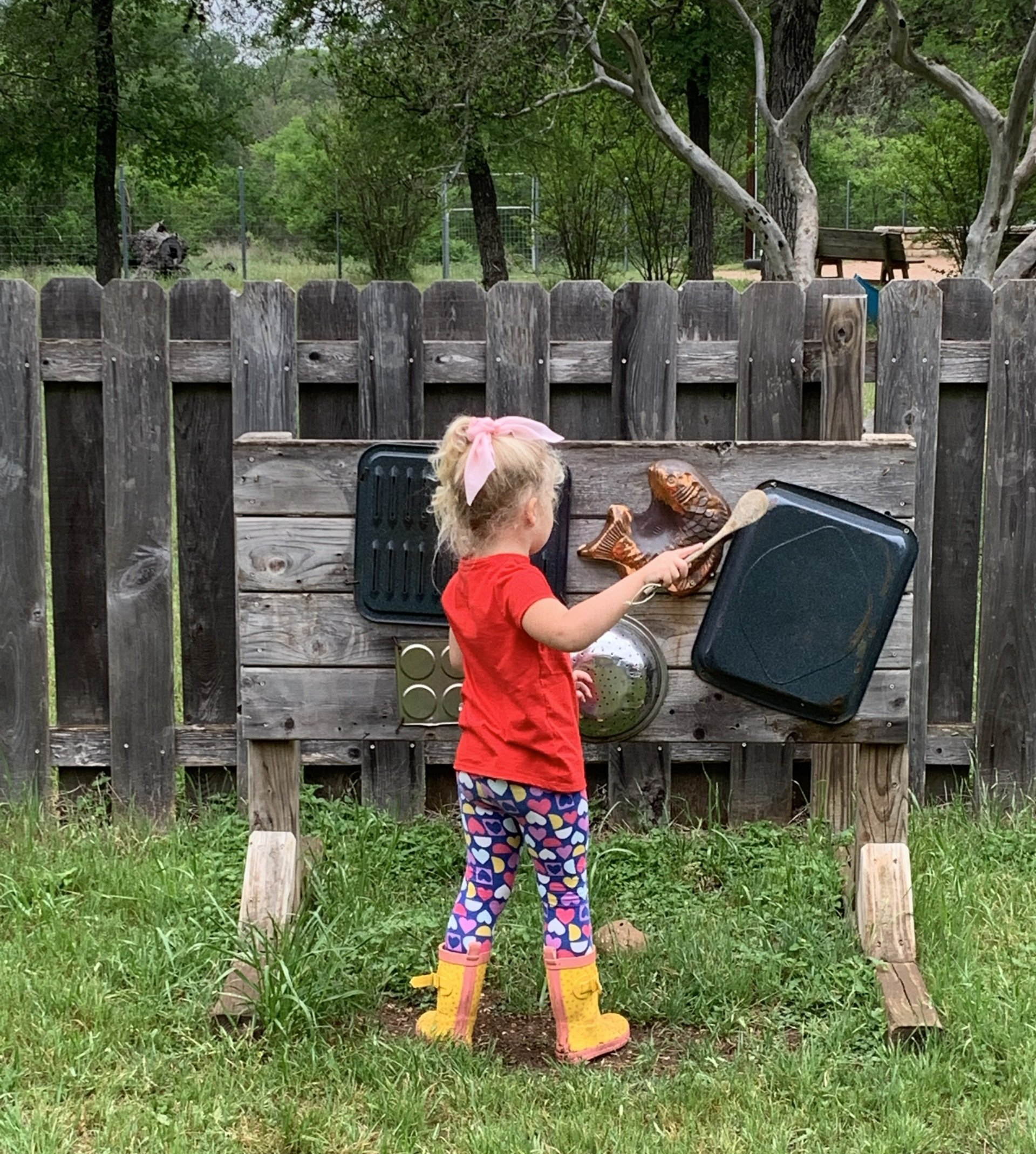 A little girl in a red shirt and yellow boots is standing in front of a wooden board.