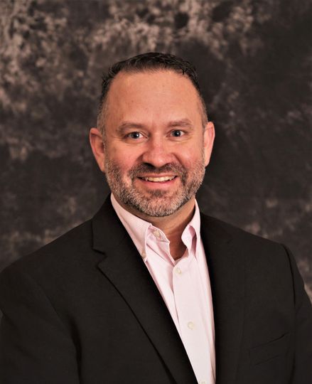 Man in black suit, pink shirt, smiling, against a mottled gray backdrop.