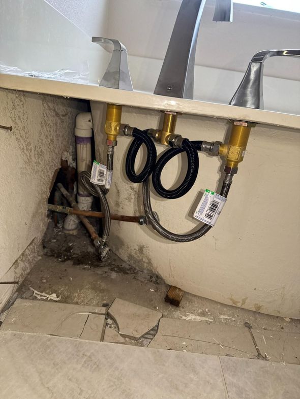 Plumbing under a bathroom sink: pipes, faucets, and flexible hoses. The wall is concrete with cracks in the floor tiles.