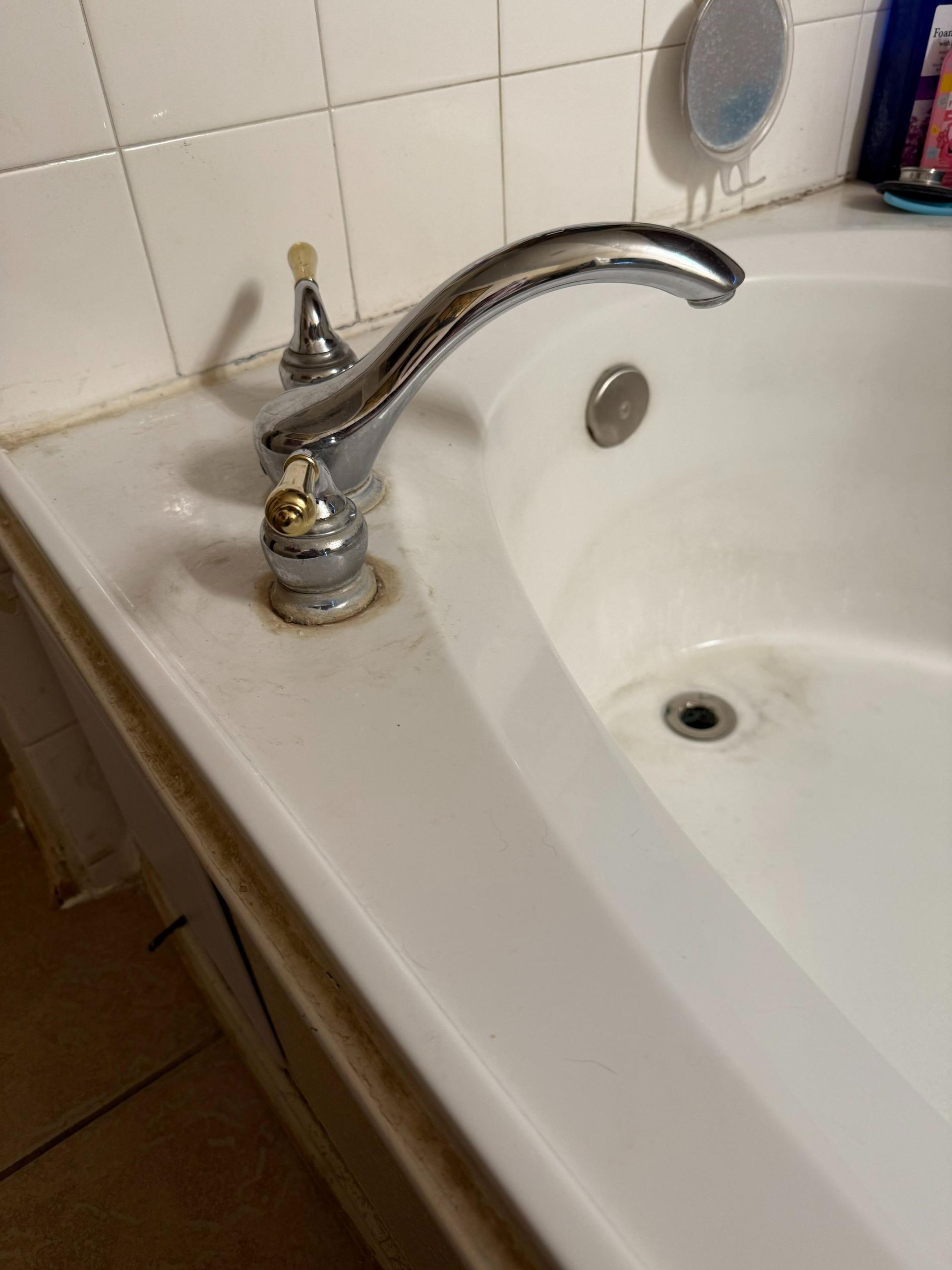 Bathtub faucet with handles, next to tub and tiled wall. Chrome fixtures, white tub, with some staining.