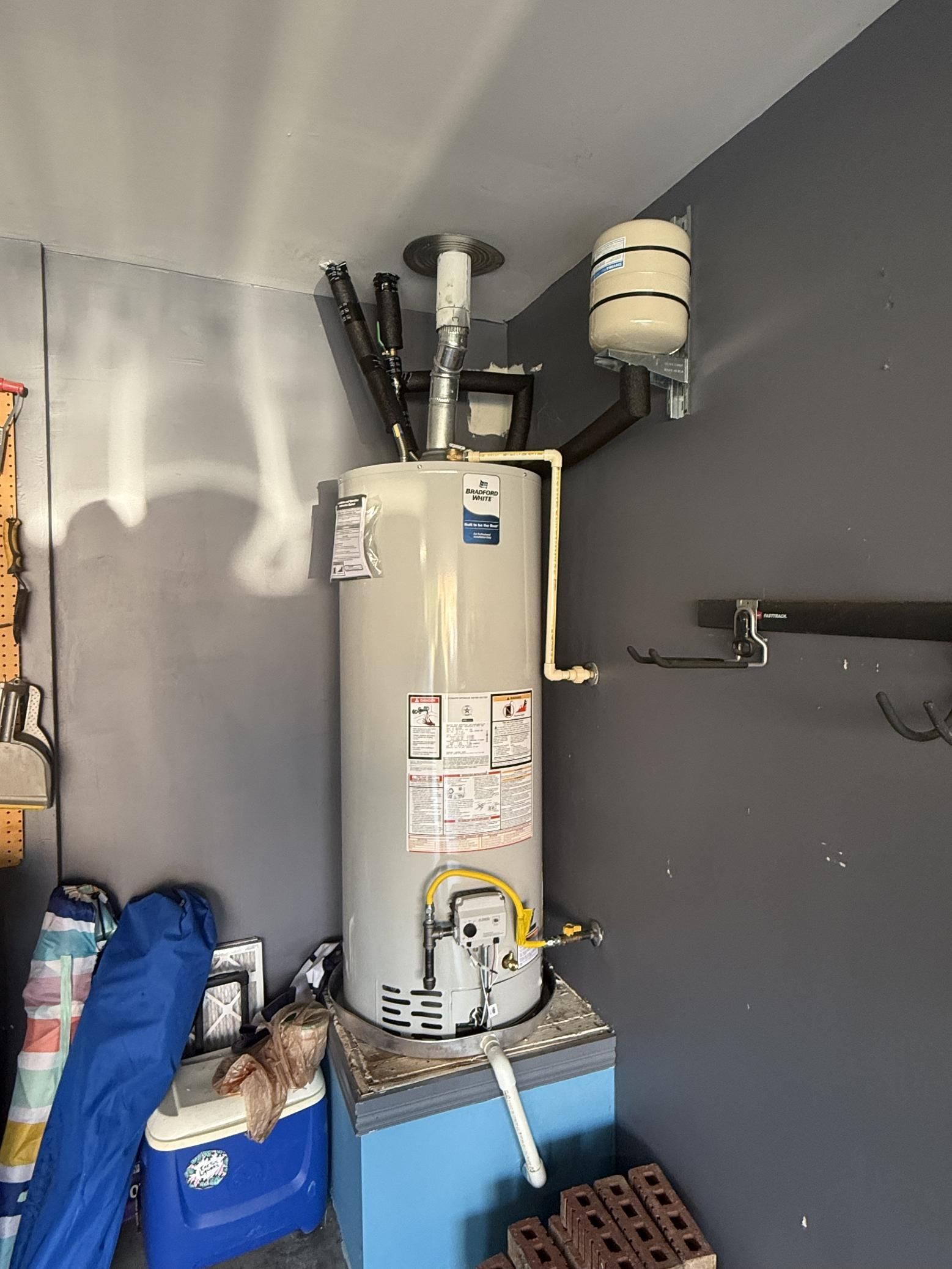 A gas water heater with attached components against a gray wall in a garage.