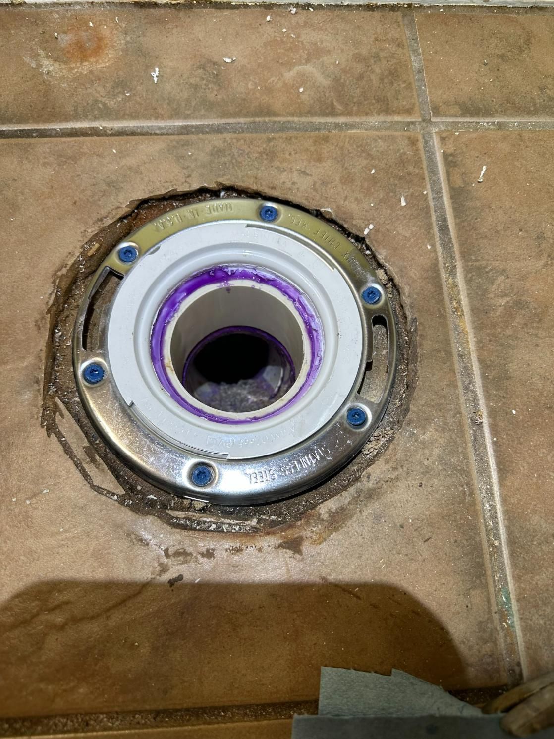 Toilet flange mounted in floor tile, showing a PVC drain pipe; purple primer visible.