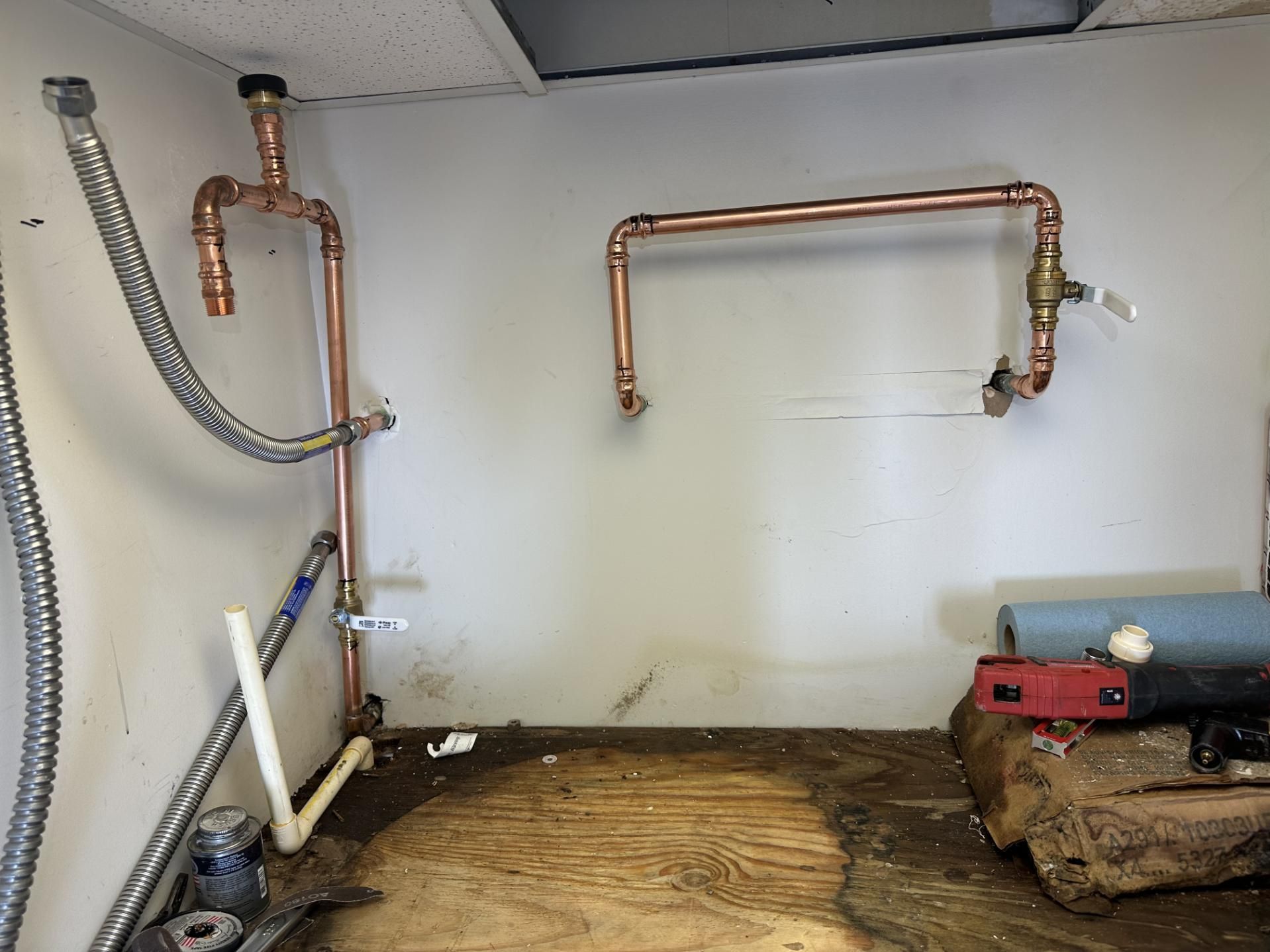 Copper pipes installed on a white wall in a room with a stained wooden floor and a drop ceiling.