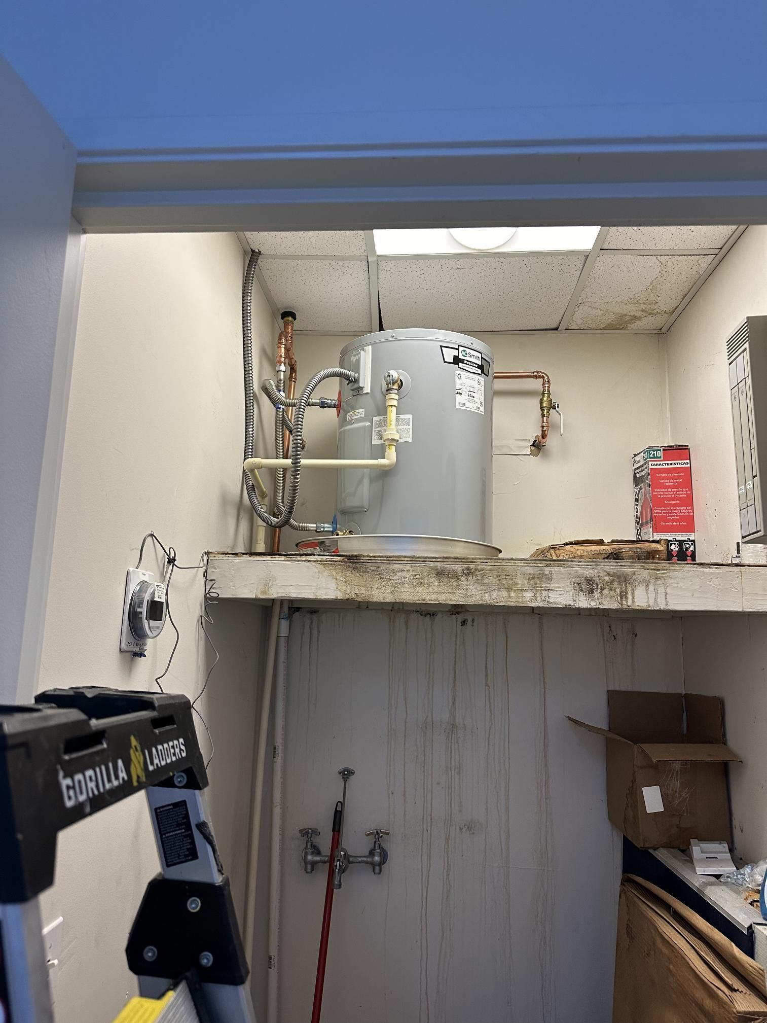 Water heater in an enclosed space with copper pipes and a partially visible ladder below.