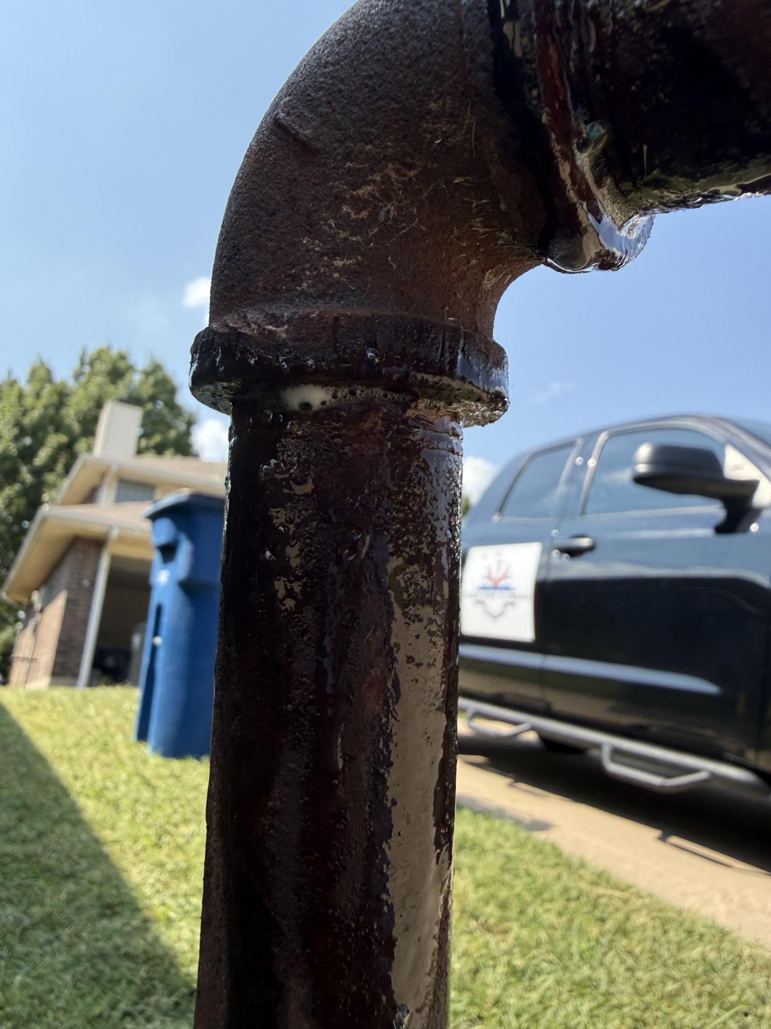 Black pipe joint leaking water, outdoors, blue sky. A vehicle, house, and trash can are in the blurred background.