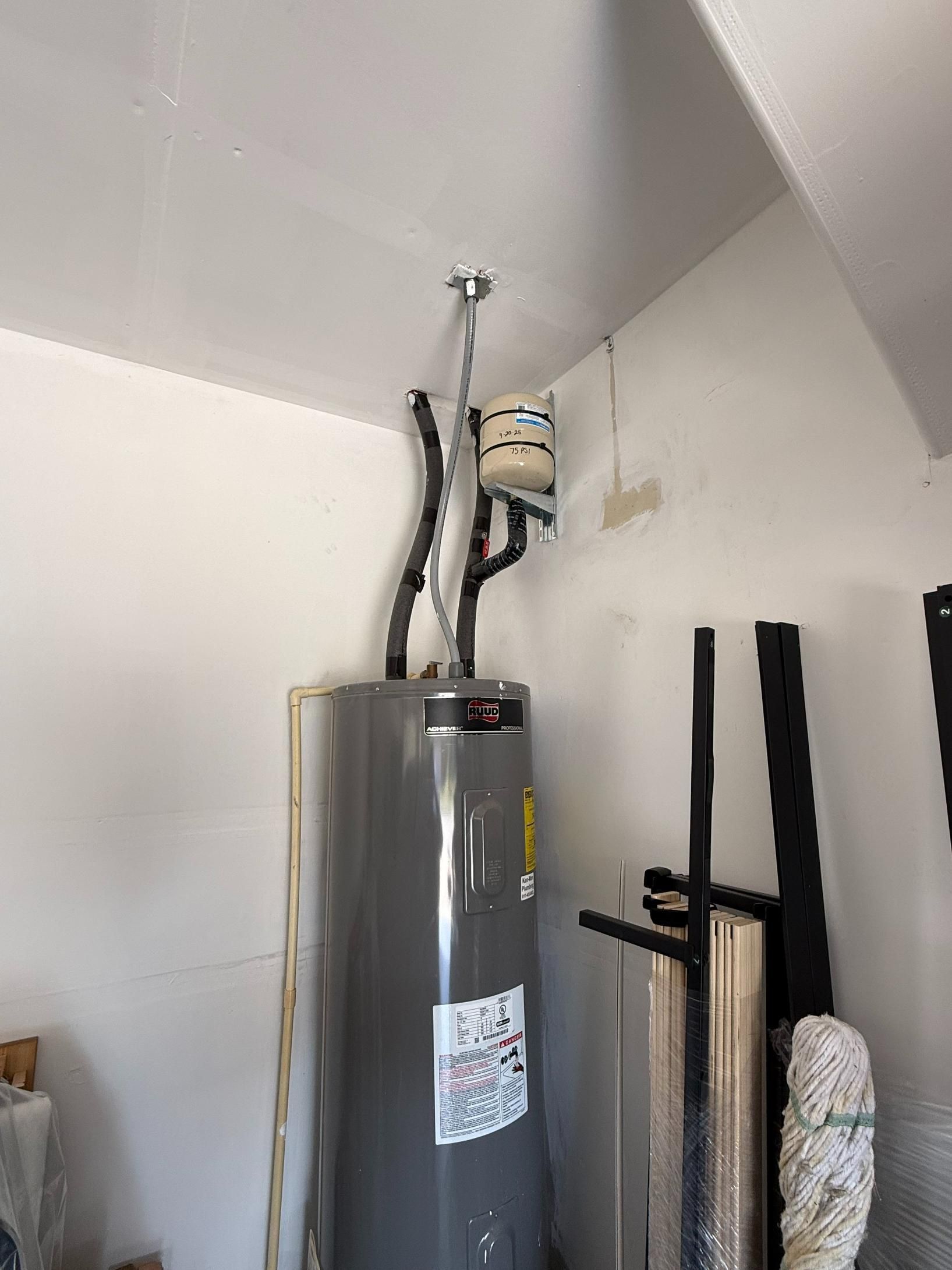 Water heater in a garage, with pipes and a white container overhead.