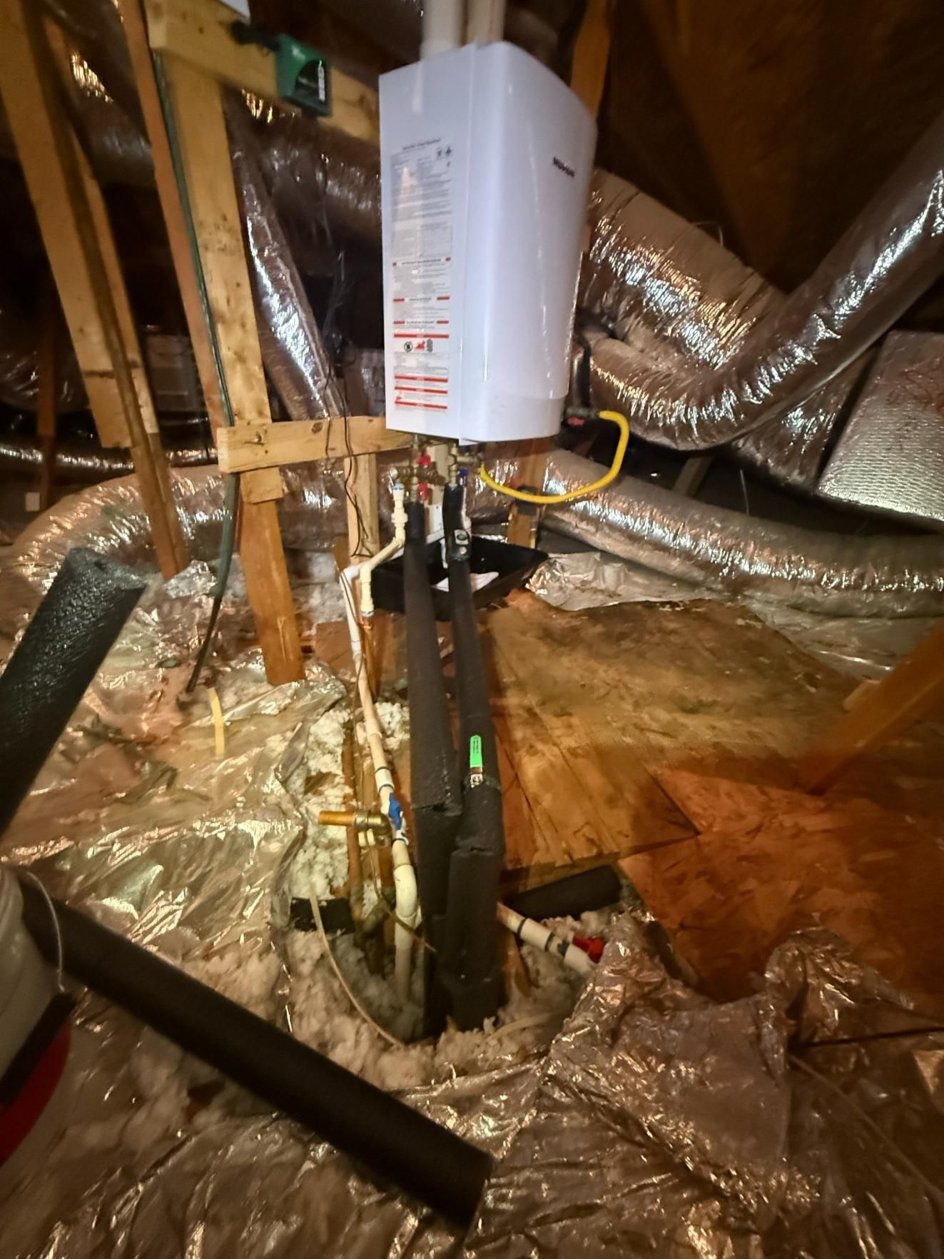 Tankless water heater installed in an attic with insulated ductwork and wood framing.