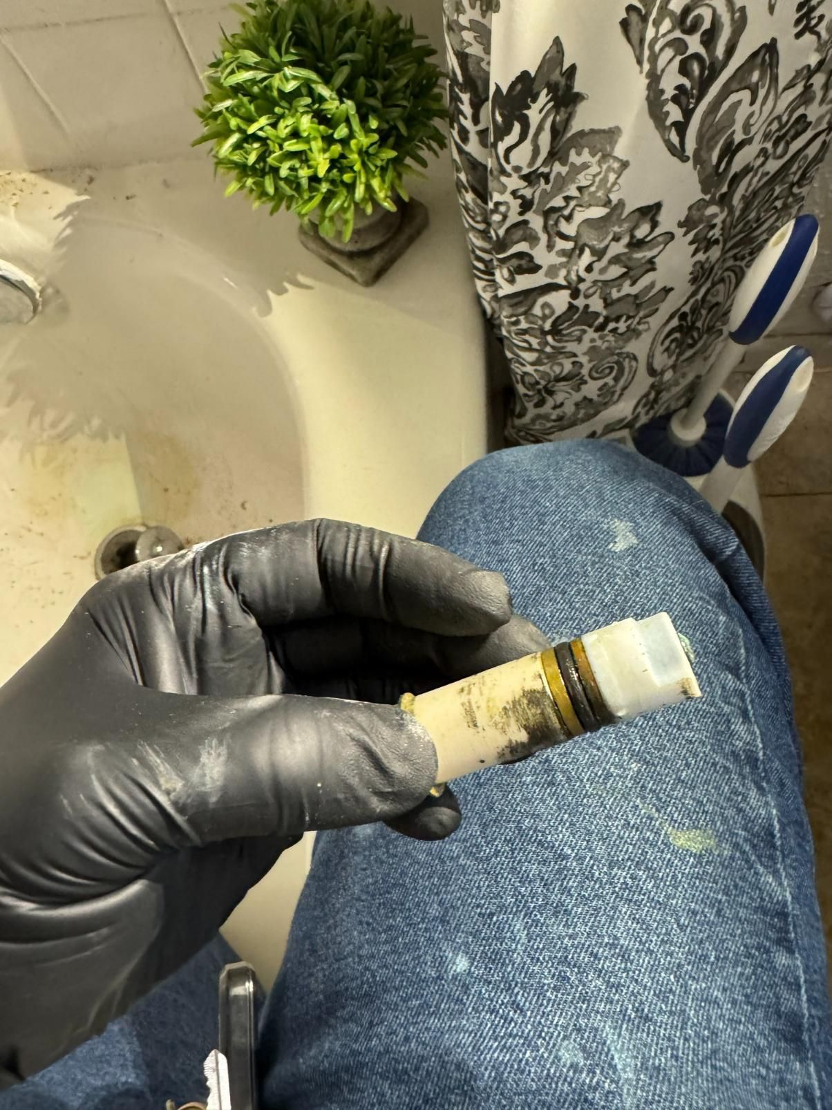 Gloved hand holding a worn cylindrical sink faucet part in a bathroom, near a sink and small plant.