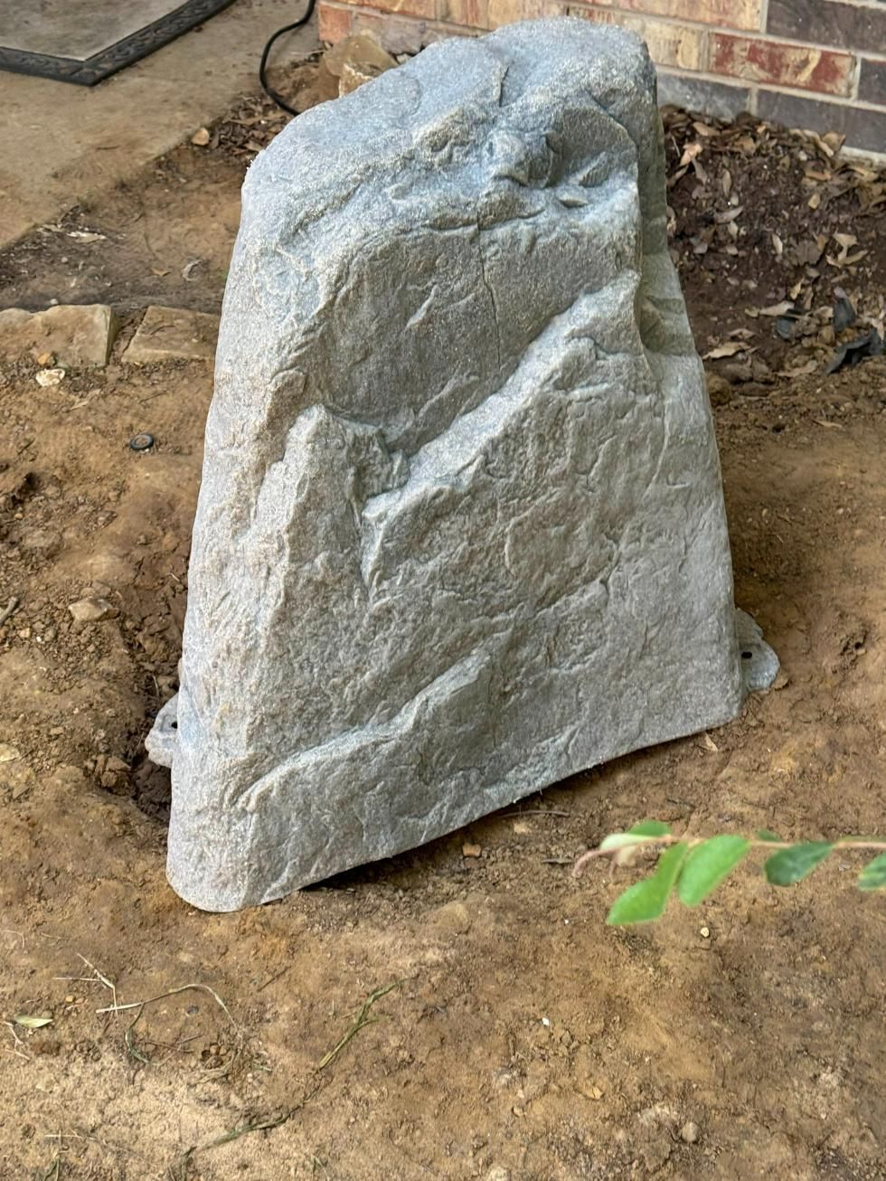 Gray rock-shaped outdoor speaker in dirt near a brick wall.