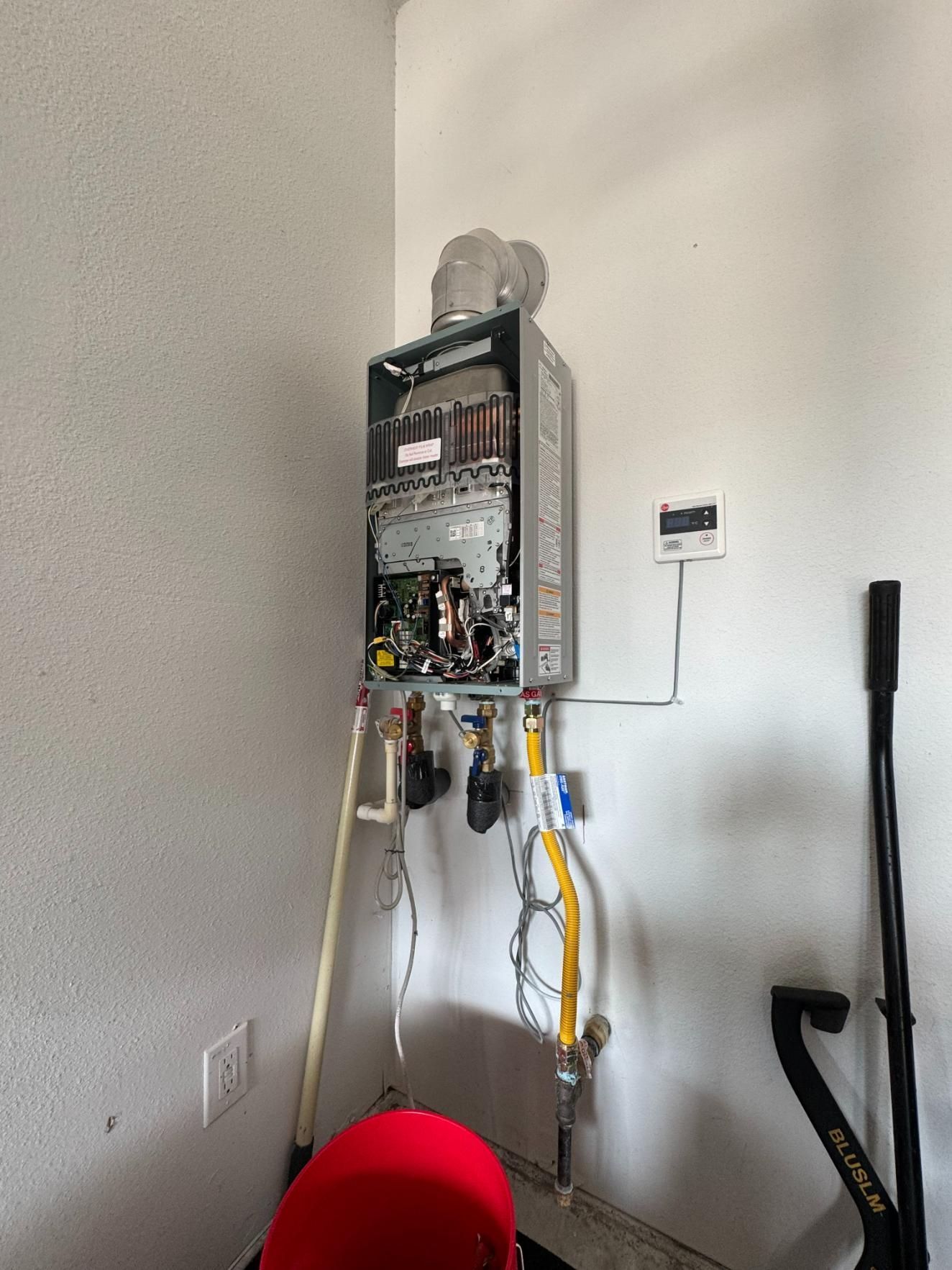 Tankless water heater installed on a white wall; pipes, wiring visible. A red object sits below.
