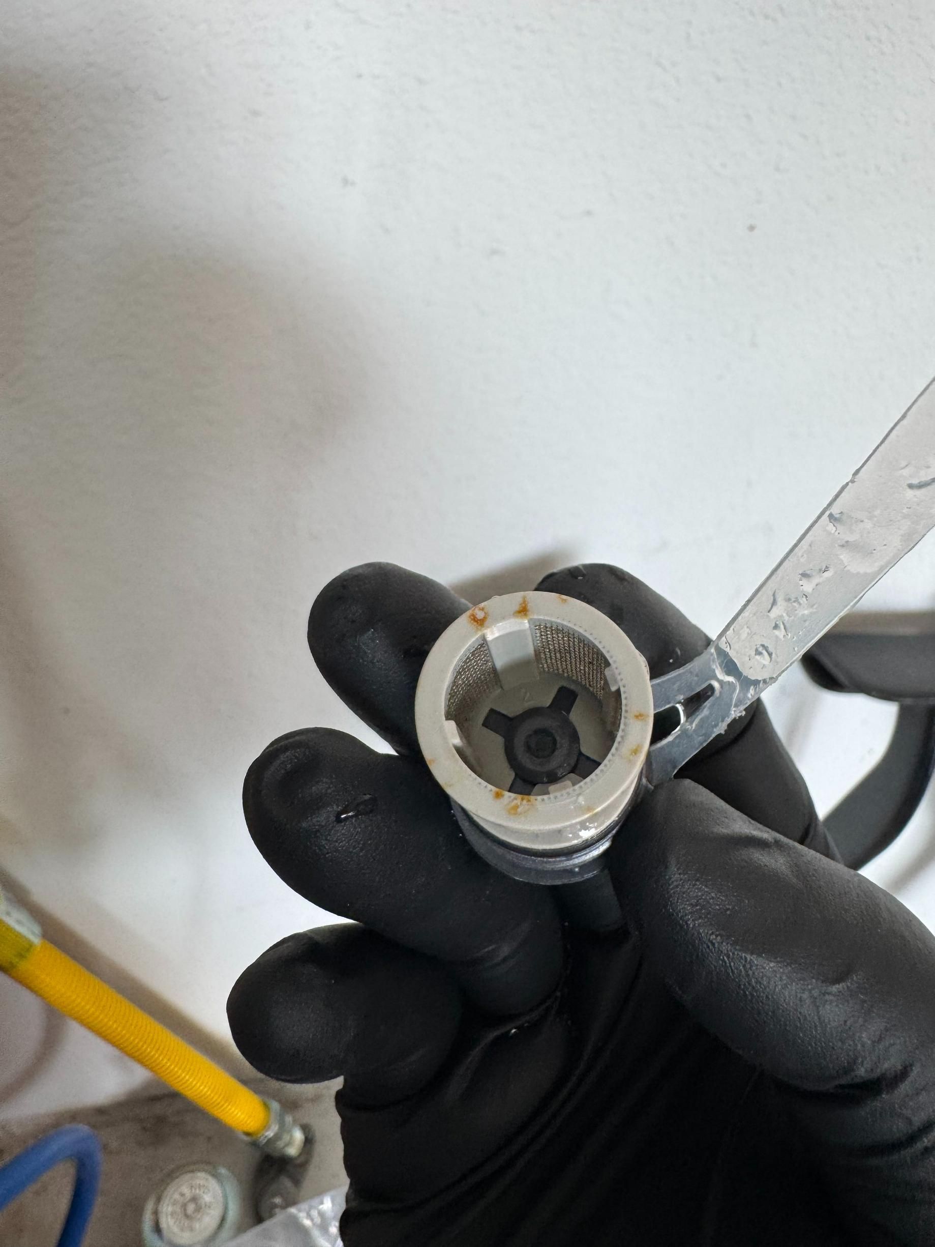 Hand in black glove holding a faucet aerator with a tool, against a white wall.