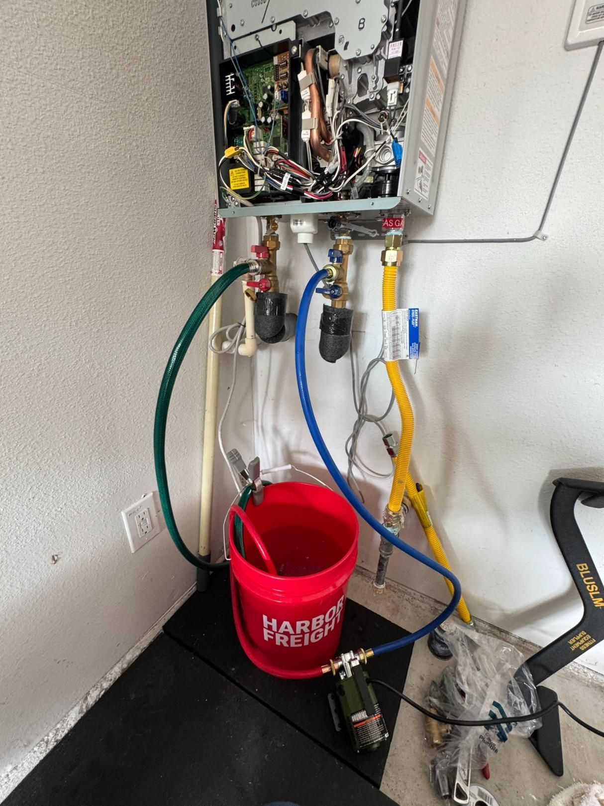 A tankless water heater with hoses and a pump connected to a red bucket, in a garage setting.