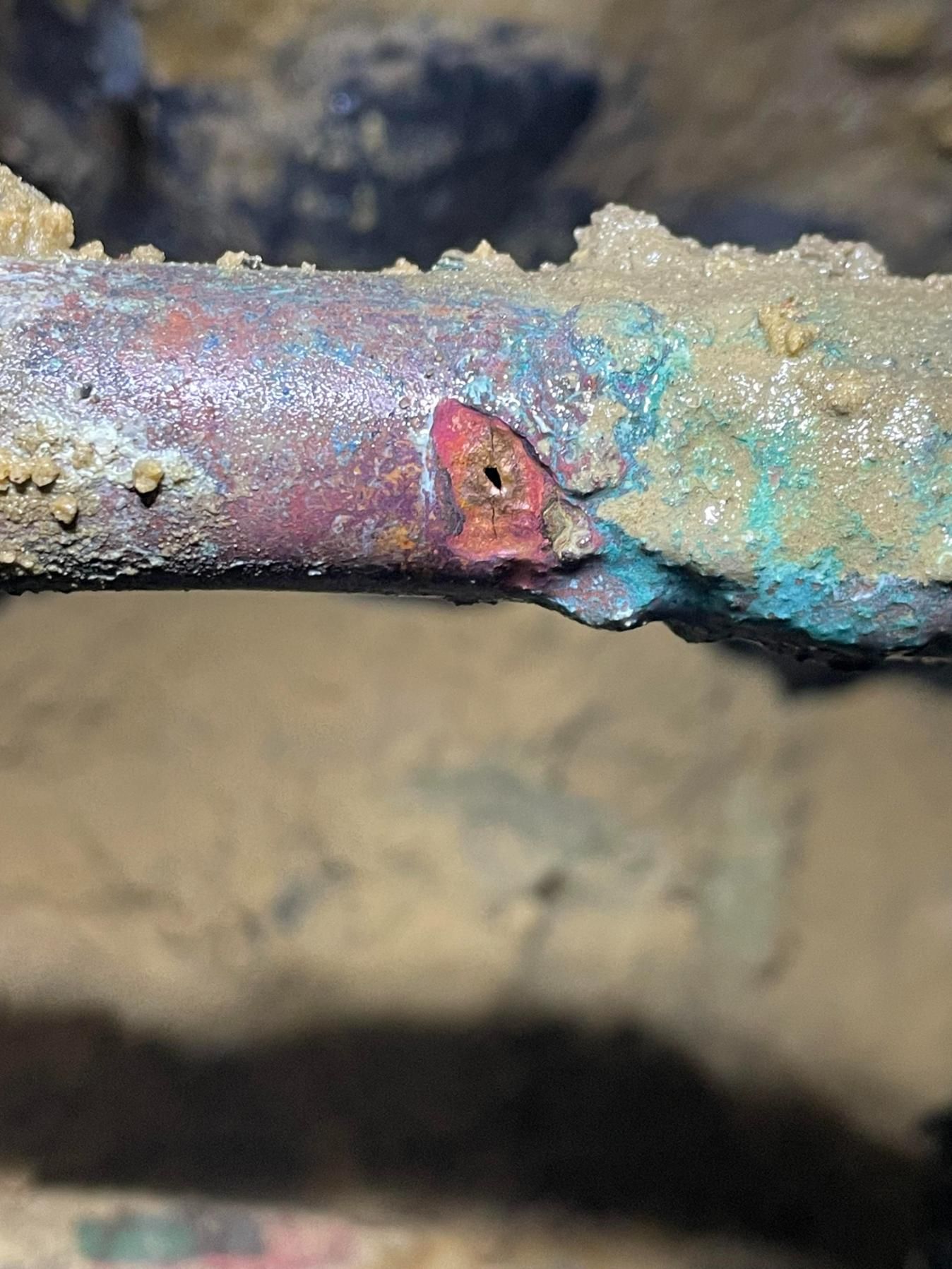 Corroded copper pipe with a hole, surrounded by green and brown residue, underground.
