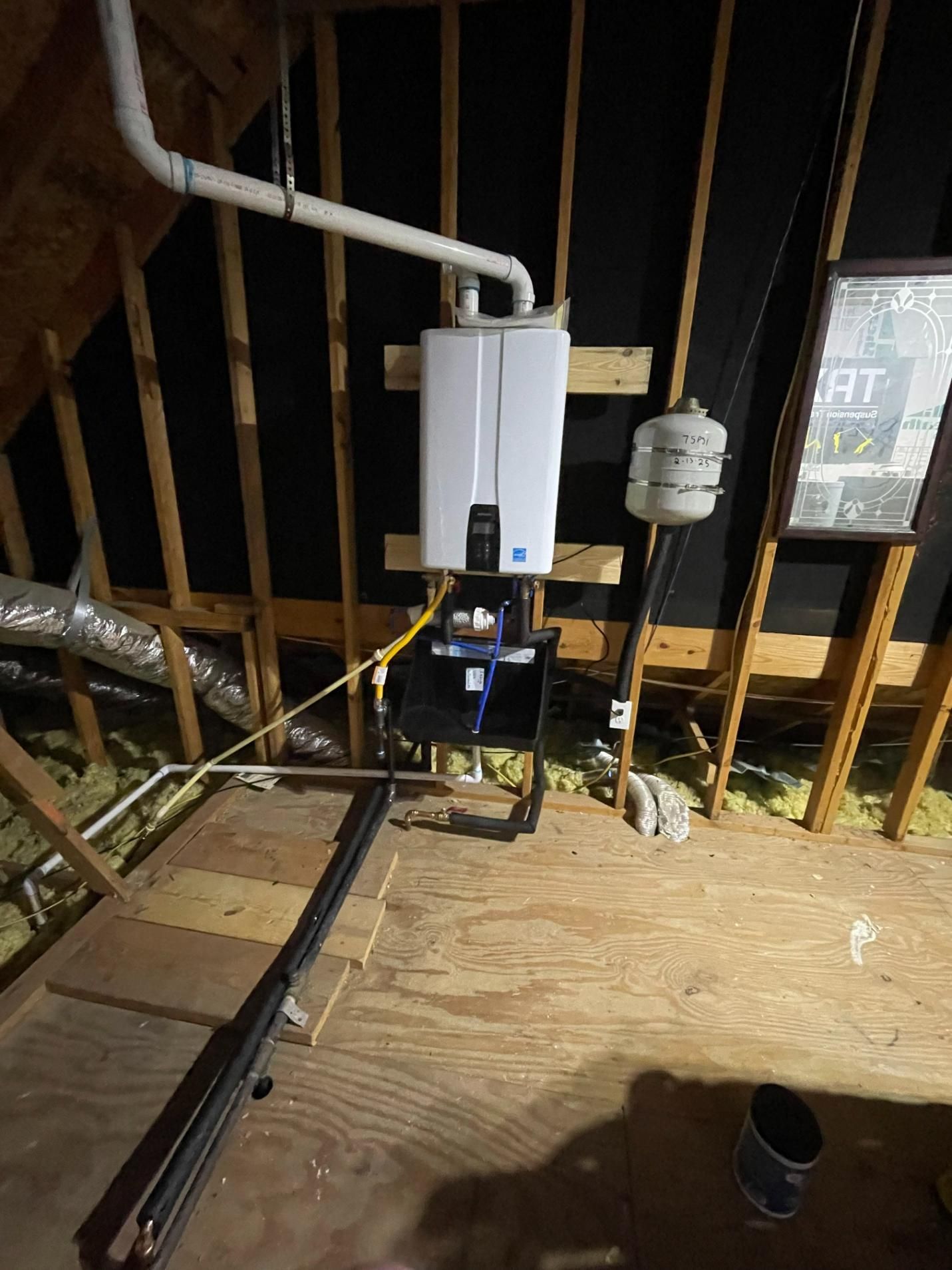 A tankless water heater installed in an attic. White pipes, a black stand, and wooden beams are visible.
