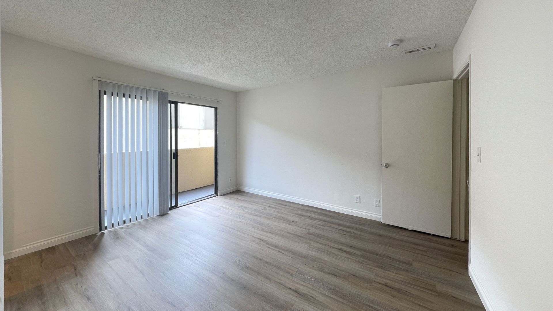 Empty apartment interior with sliding glass door and vertical blinds, light wood-look floors, and white walls.