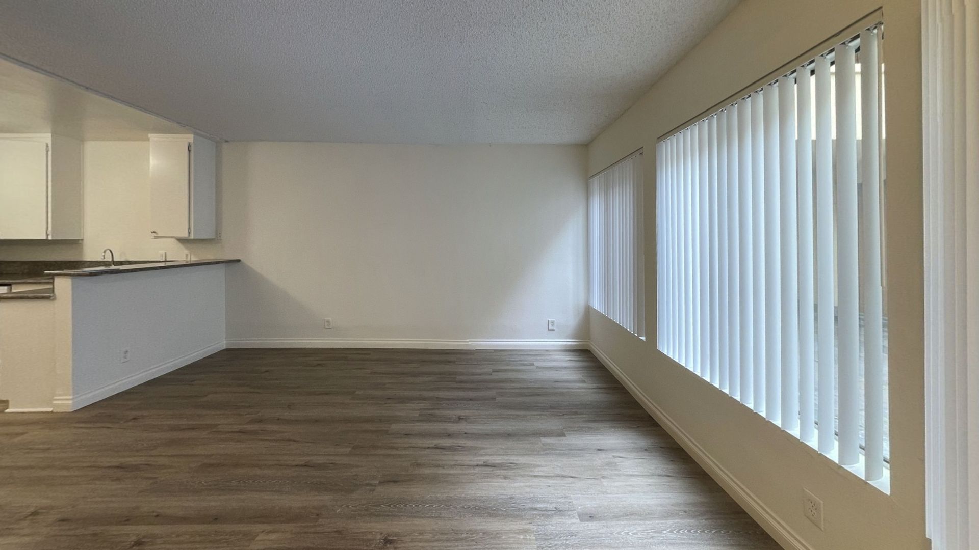 Empty apartment living space with wood-look floor, white walls, and vertical blinds.