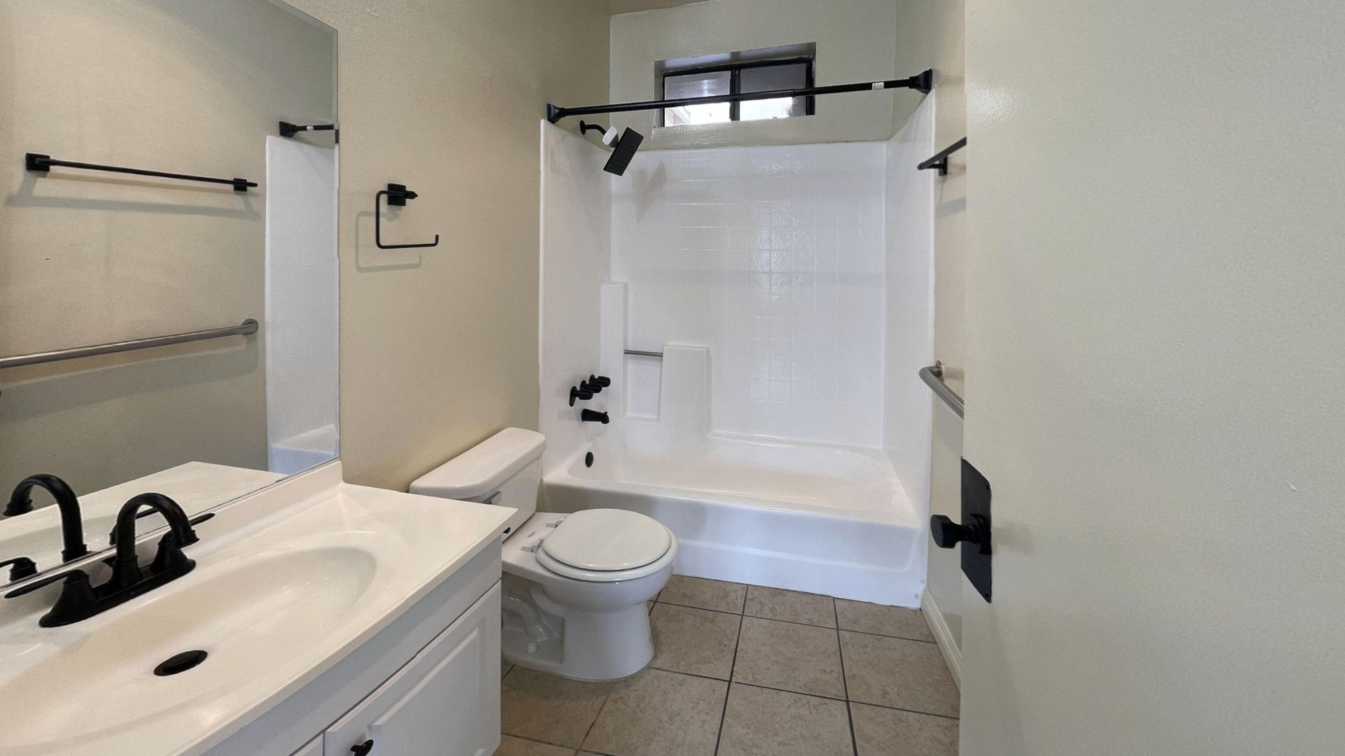 Bathroom with a white shower, toilet, and sink. Black hardware and tan floor tiles.