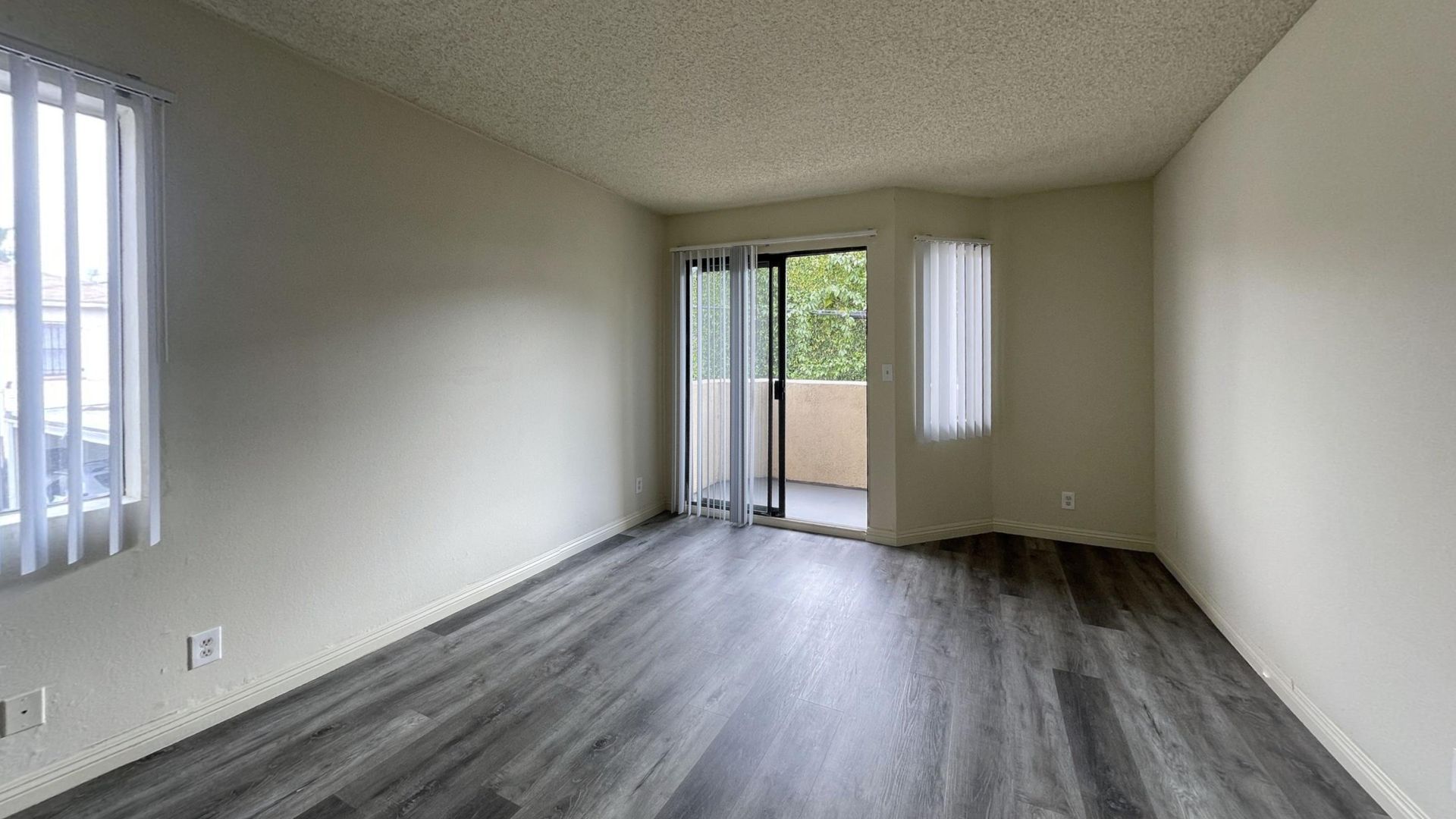 Empty room with gray wood-look flooring, white walls, two windows with blinds, and a sliding glass door to a balcony.
