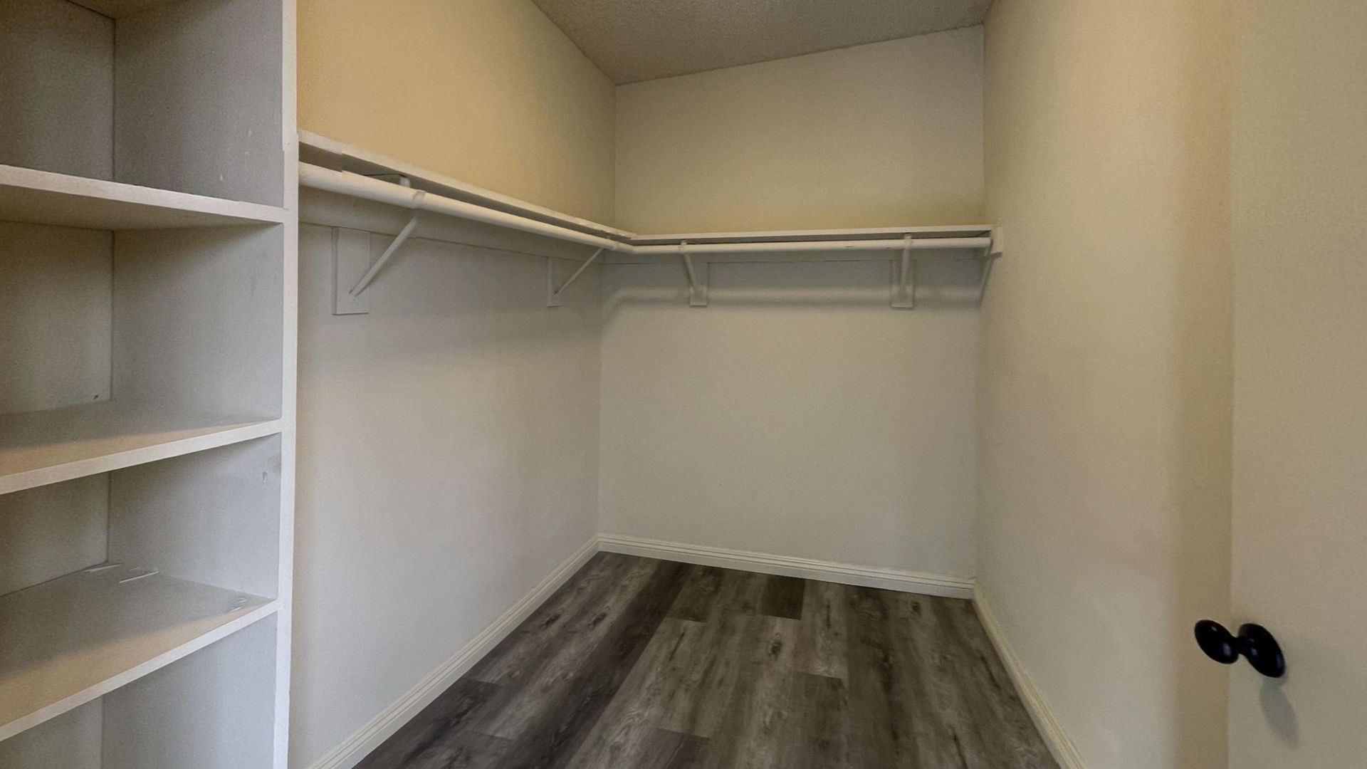 Empty walk-in closet with shelves and hanging rods. Gray wood-look floor, beige walls.