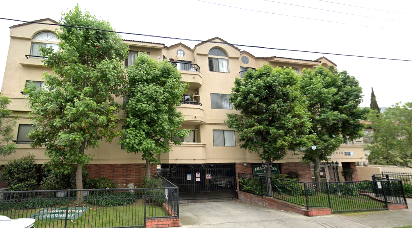 Apartment building with beige exterior, three stories, and trees in front of a gated entrance.