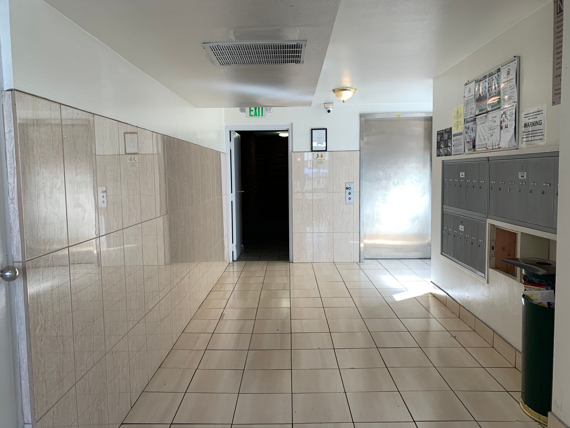 Hallway with tiled walls and floor, mailboxes, and an open doorway leading to a dark space.